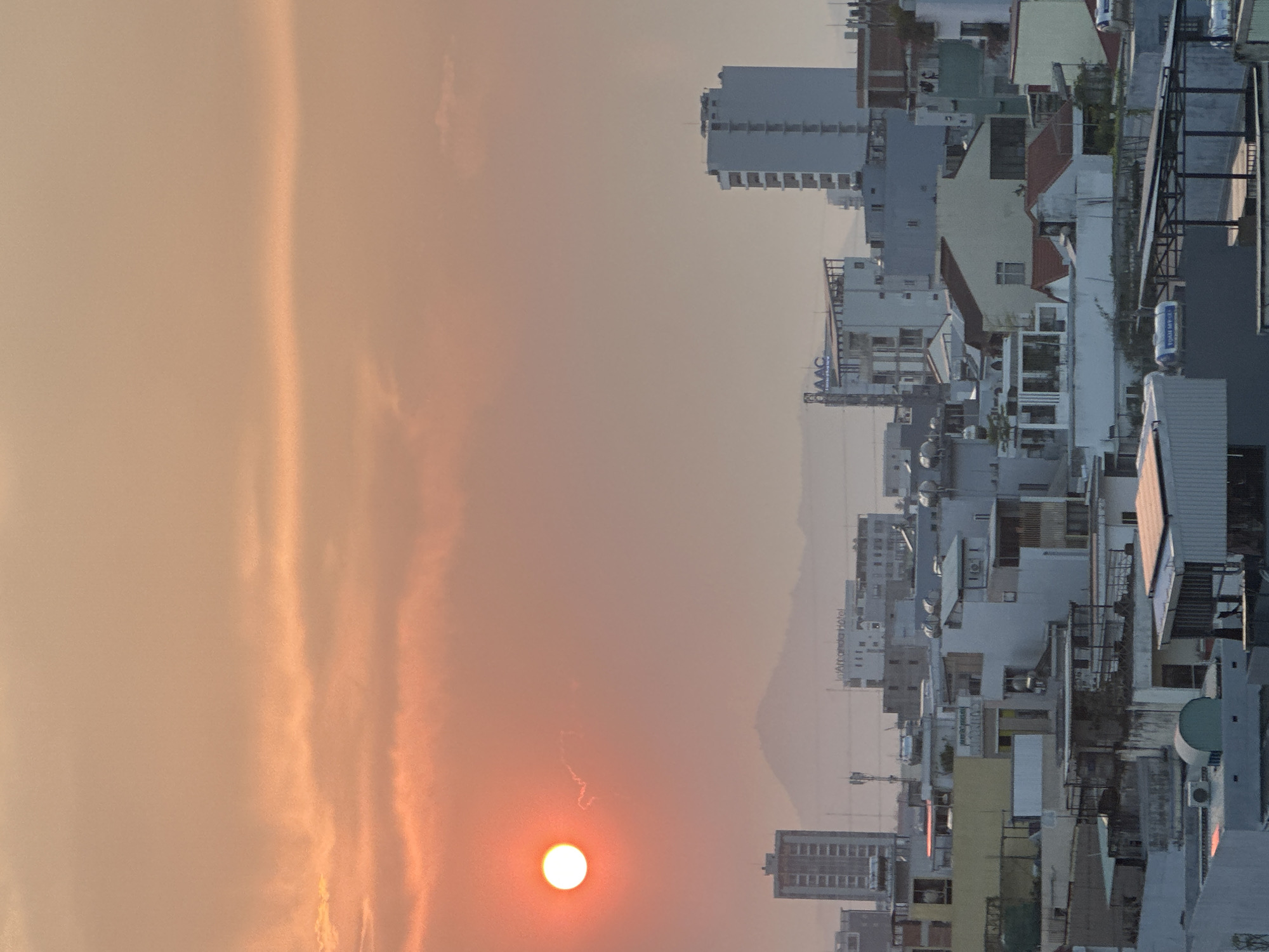 An outdoor view oriented vertically shows a cityscape at sunrise. The sun is visible at the bottom left corner, with orange hues dominating the sky. Silhouetted mountains are faintly visible in the background. Buildings of various heights and designs line the horizon on the right. The city is Đà Nẵng, Vietnam.