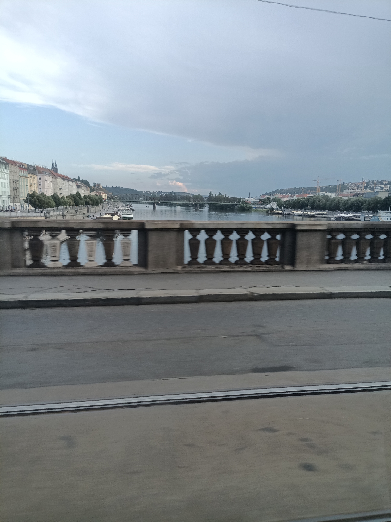 The image captures a view from a bridge in Prague, Czechia, looking out over the Vltava River. In the foreground, the ornate stone balustrade of the bridge is visible, with its repeating pattern of decorative pillars. Beyond the balustrade, the river stretches out, reflecting the overcast sky. On the far bank, a line of historic buildings with varied architectural styles can be seen, interspersed with trees. Further in the distance, a larger bridge crosses the river, and the city sprawls up the hills, with structures and construction cranes dotting the skyline. The foreground also shows a section of asphalt roadway and what appear to be tram tracks, suggesting the vantage point is from a moving vehicle or a pedestrian walkway on the bridge. The overall atmosphere is one of a city scene during daylight hours under cloudy skies.