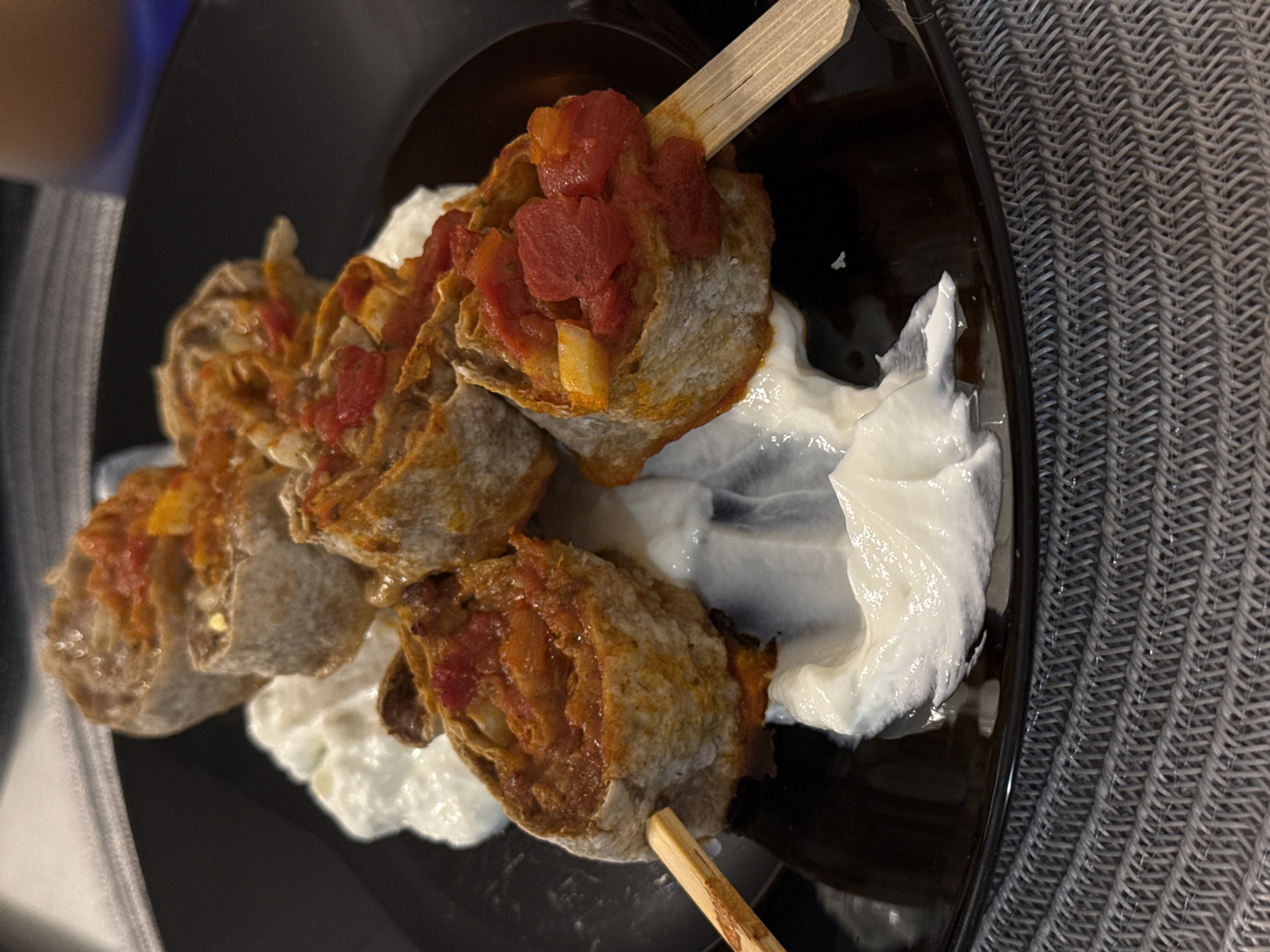 A close-up, slightly rotated overhead shot shows a black plate containing what appears to be a Bulgarian dish, possibly "Shishcheta" (skewered meat), served on a bed of white sauce (likely yogurt or garlic sauce). The skewers have pieces of toasted bread rolled and held together with wooden sticks. The rolls are topped with a tomato-based sauce. The plate sits on a gray woven surface. The location cannot be inferred from the image. Sofia, Bulgaria, is not identifiable from the image alone.