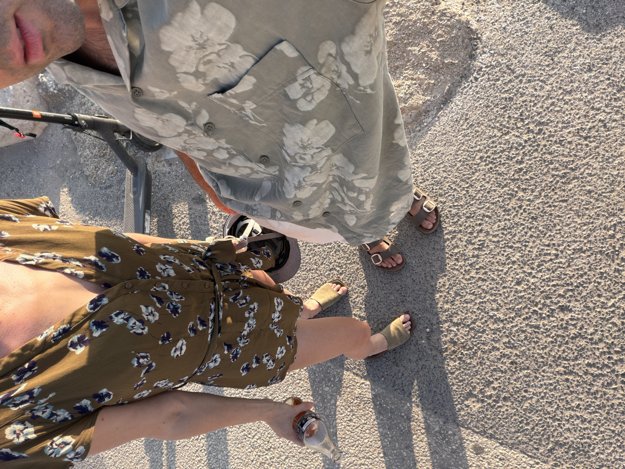 The image shows a close-up, high-angle view of two people standing on a paved surface in Tel Aviv, Israel. The person on the left is wearing a green floral-print romper and olive-green sandals, holding a glass bottle. The person on the right is wearing a light-colored floral-print shirt and brown sandals. An electric scooter is visible in the background. The lighting suggests it is either early morning or late afternoon. The ground appears to be a textured asphalt or concrete surface, with shadows cast by the individuals.