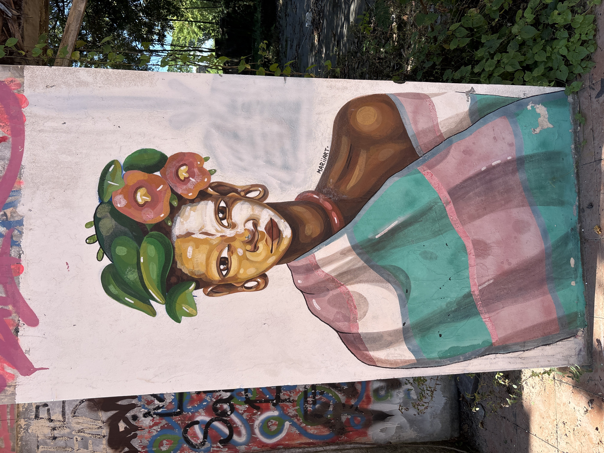 The image shows a mural of a woman on a concrete wall, likely an outdoor setting. The woman is depicted with dark brown skin, adorned with greenery and flowers in her hair. She wears a striped garment in shades of green, pink, and white. The mural is signed "MAR11ART". The wall also has other graffiti markings beneath the mural. The top of the wall is partially covered by foliage. The location is assumed to be in Denpasar, Indonesia.