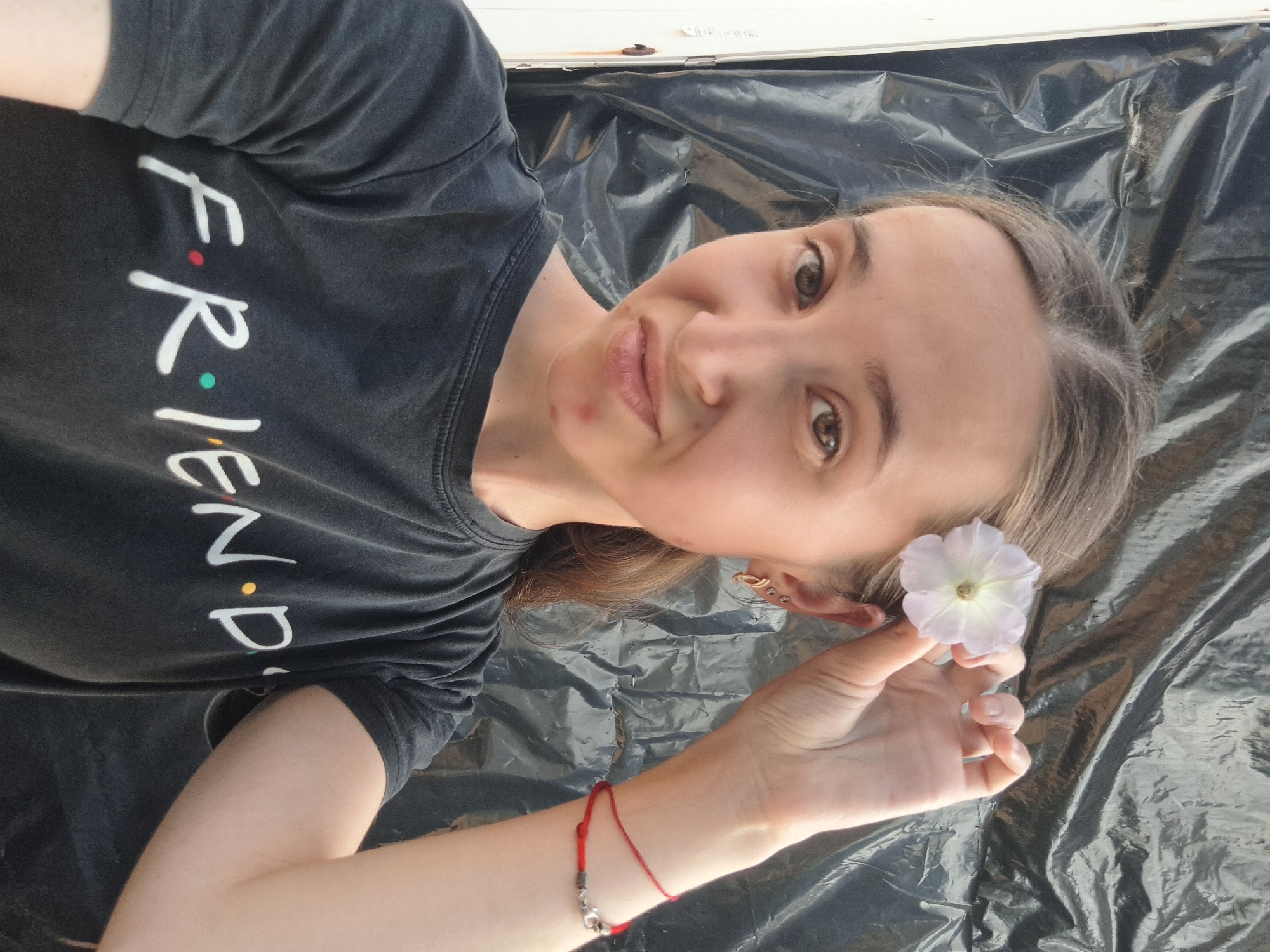 A young woman is taking a selfie in front of a dark, crinkled background, possibly tarpaulin, set against a white vertical structure. She is wearing a black t-shirt with the word "FRIENDS" printed on it in white lettering, with colored dots replacing the letter 'i'. She has a light pink flower tucked behind her right ear, held in place by her fingers. She also wears a red string bracelet on her right wrist and earrings in her left ear. Her facial expression is playful, with a slight pout. The lighting suggests an outdoor setting, and the overall composition is a close-up portrait. There are no discernible landmarks to pinpoint the location within Vypasne, Ukraine.