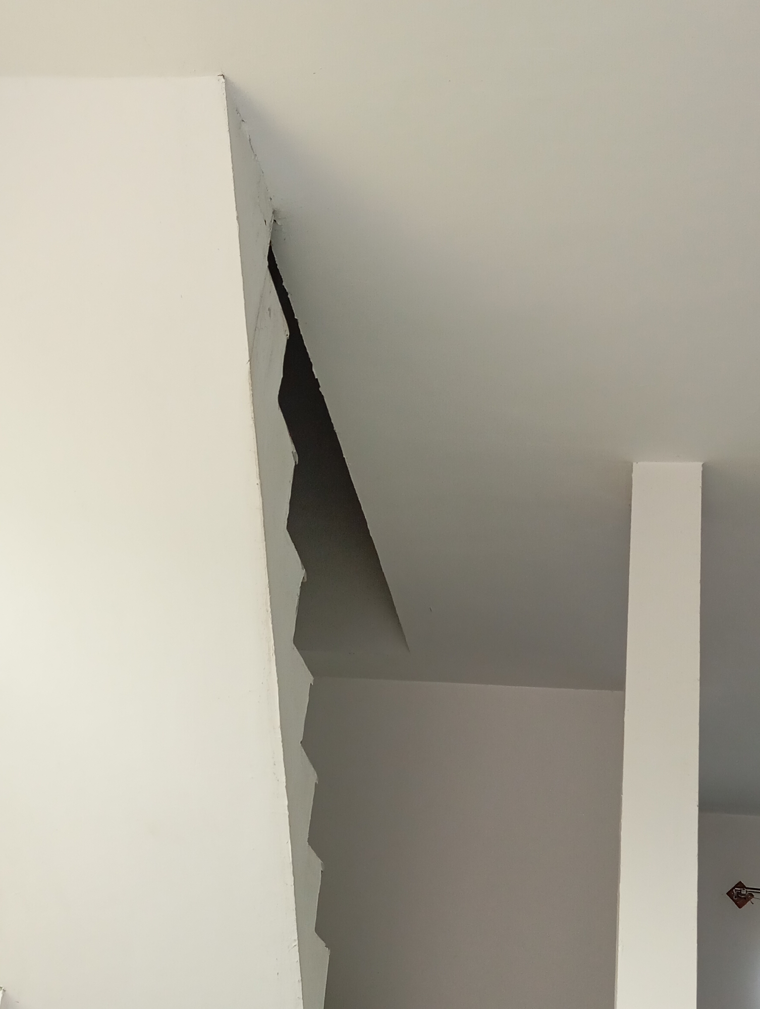 A wide-angle, close-up, eye-level shot shows a white ceiling with a large, jagged crack in the corner. The crack creates a sharp, zigzagging opening into a dark void. The surface of the ceiling appears to be drywall. To the right of the crack, a white rectangular column extends vertically from floor to ceiling, out of frame. The wall on the left is also white. The scene is indoors, with no indication of location other than a white ceiling, a column, and a crack.