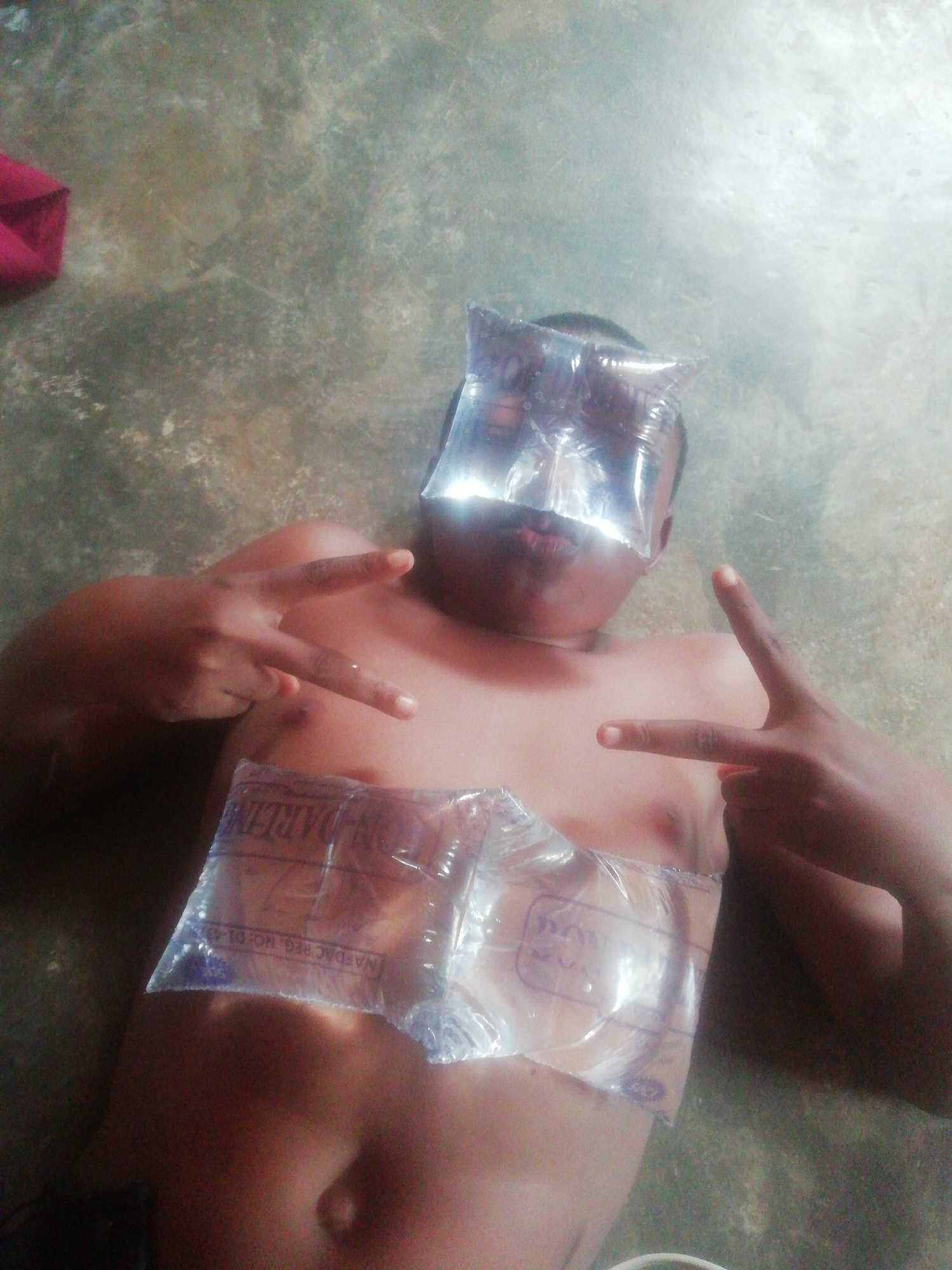A person is lying on their back on a floor. Two small bags of water, one on their eyes and the other on their chest, obscure portions of the subject. The individual is making a peace sign with both hands. Part of a red cloth is visible in the upper left corner of the image.