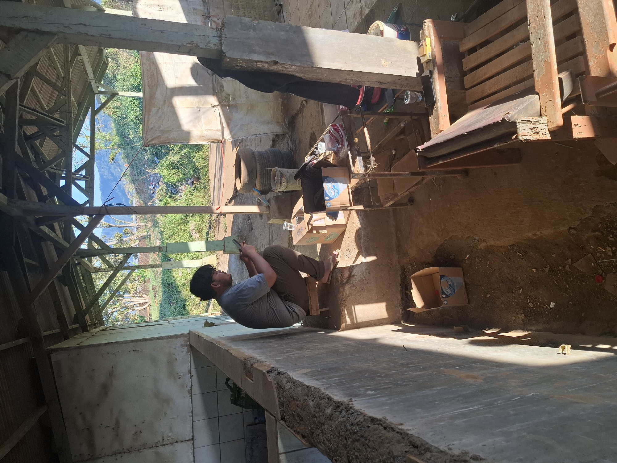 A young man is sitting on a low wooden stool, facing right and looking down at his smartphone. He is wearing a gray t-shirt and dark pants. The man is seated in a covered, open-air structure with a corrugated metal roof supported by wooden beams and concrete pillars. To his left is a concrete wall, and to his right, there is a table with various items including boxes, containers, and a glass of water. In the background, the scene opens to a rural landscape with trees and hills under a clear blue sky. The ground within the structure is dirt and debris, with a few scattered objects including an empty cardboard box and cigarette butts. The structure appears to be utilitarian, possibly a workshop or storage area, located in a rural setting.