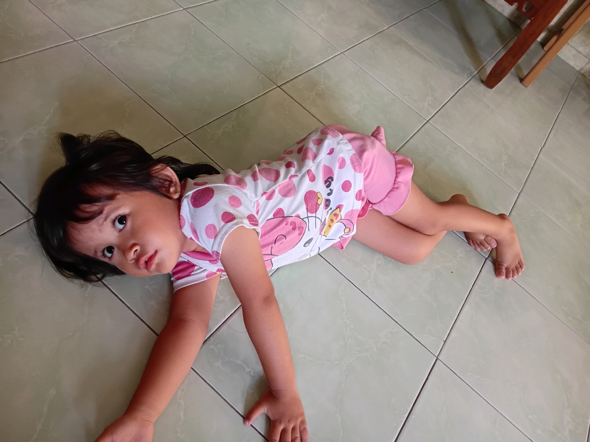 A young girl with dark hair lies on her side on a tiled floor. She is wearing a white and pink polka-dotted outfit with ruffles on the bottom. Her eyes are open and she is looking towards the camera with a slightly quizzical expression. The room appears to be indoors, with a light-colored, tiled floor. To the right of the frame, a portion of a wooden chair is visible. The lighting suggests it could be daytime. The overall impression is a candid snapshot of a child relaxing or playing on the floor. There is no visible text in the image.