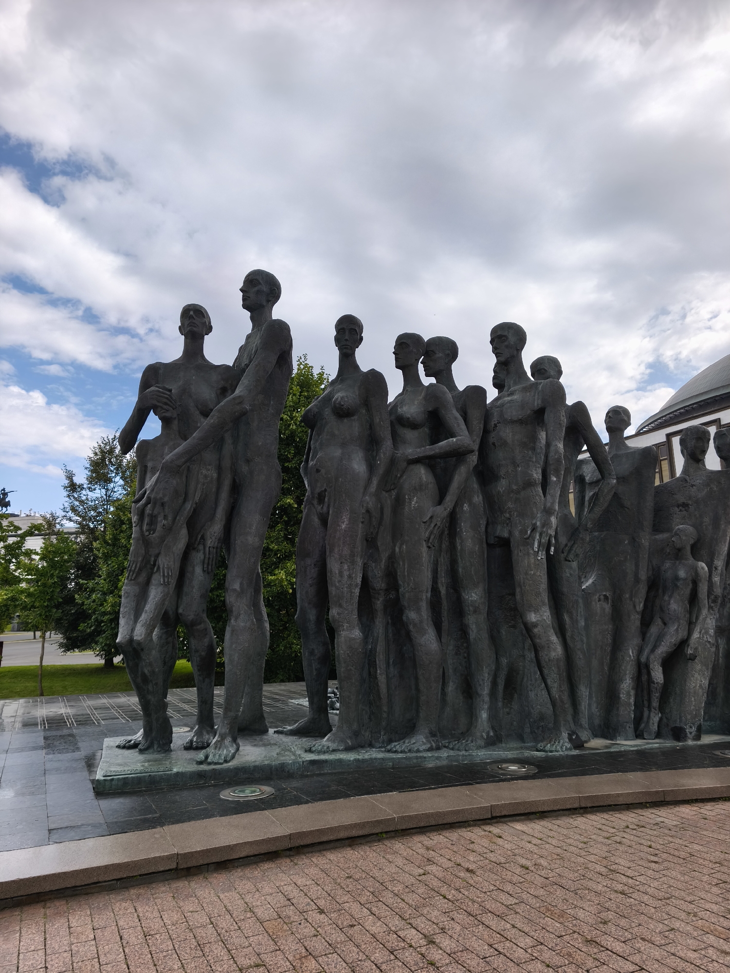 This is a daytime, exterior, eye-level, medium-wide shot of the "Temple of the Unknown Soldier" monument in Moscow, Russia. The monument, a large bronze sculpture of a procession of figures, is positioned in the foreground, with a cloudy sky and trees visible in the background. The figures are depicted in various poses, some standing, others huddled together, with an abstract and somewhat somber aesthetic. The ground in front of the monument is paved with reddish-brown bricks, transitioning to a darker, polished stone base for the sculpture itself. The overall impression is one of solemnity and remembrance, with the figures suggesting a collective journey or shared experience.