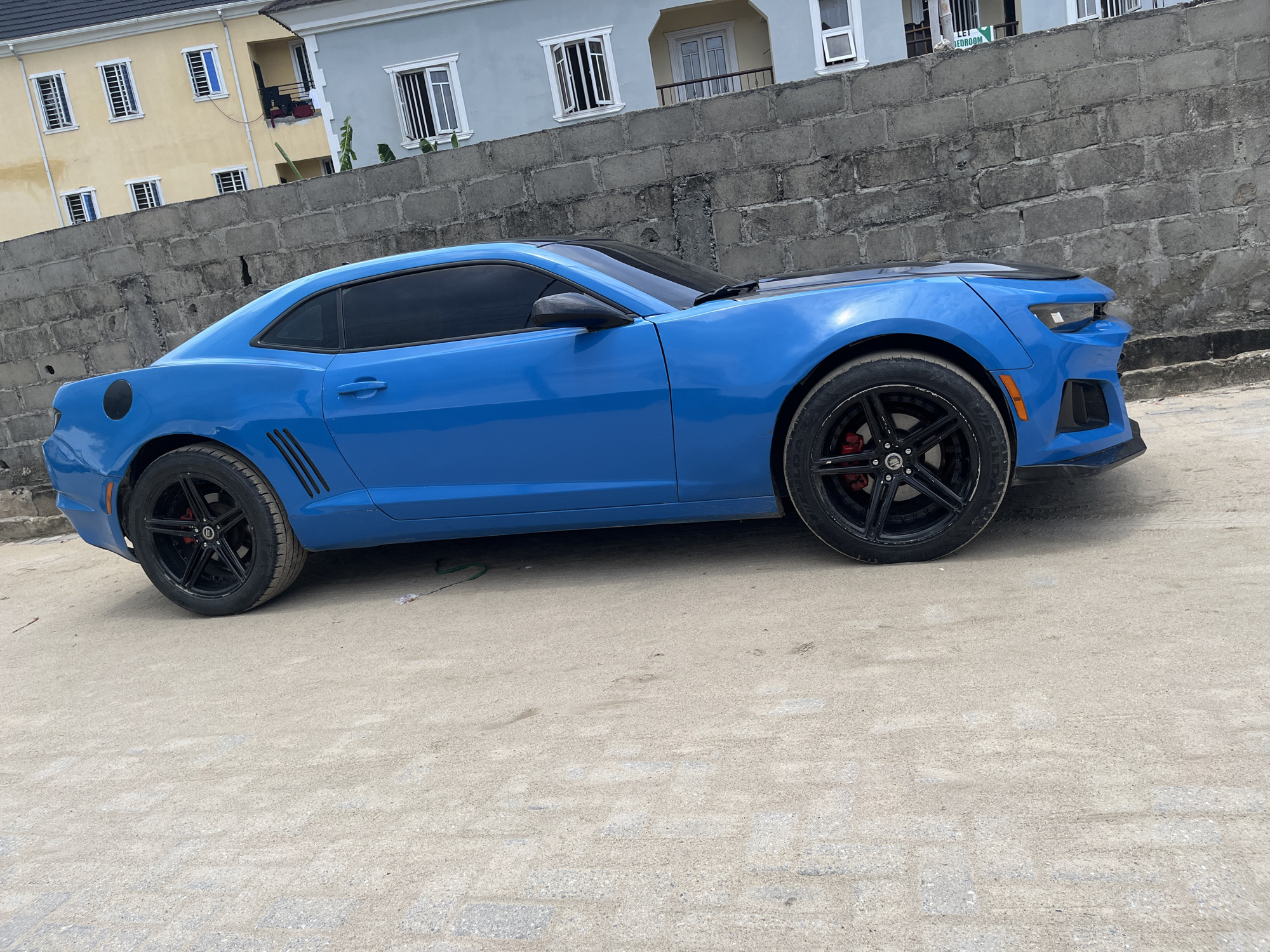 The image captures a vibrant blue Chevrolet Camaro sports car parked on a light-colored, sandy or dirt road. The car is the primary subject, featuring black multi-spoke wheels with visible red brake calipers, a contrasting black hood, and darkly tinted windows. A circular black fuel cap is located on the rear quarter panel.
In the midground, behind the car, is a sturdy concrete block wall. Beyond this wall, two multi-story buildings are visible. The building on the left is painted a pastel yellow, while the one on the right is light grey or blue. Both buildings feature multiple windows, some with white frames and vertical security bars, and the grey building includes balconies.
The scene appears to be set outdoors during bright daylight, suggesting clear and sunny weather. There is no visible human activity or movement; the car is stationary. The architecture and environmental elements are consistent with a residential or mixed-use area, aligning with the location context of Iranla, Nigeria. Partial text "LET BEDROOM" is discernible on a sign in the background associated with the grey building.