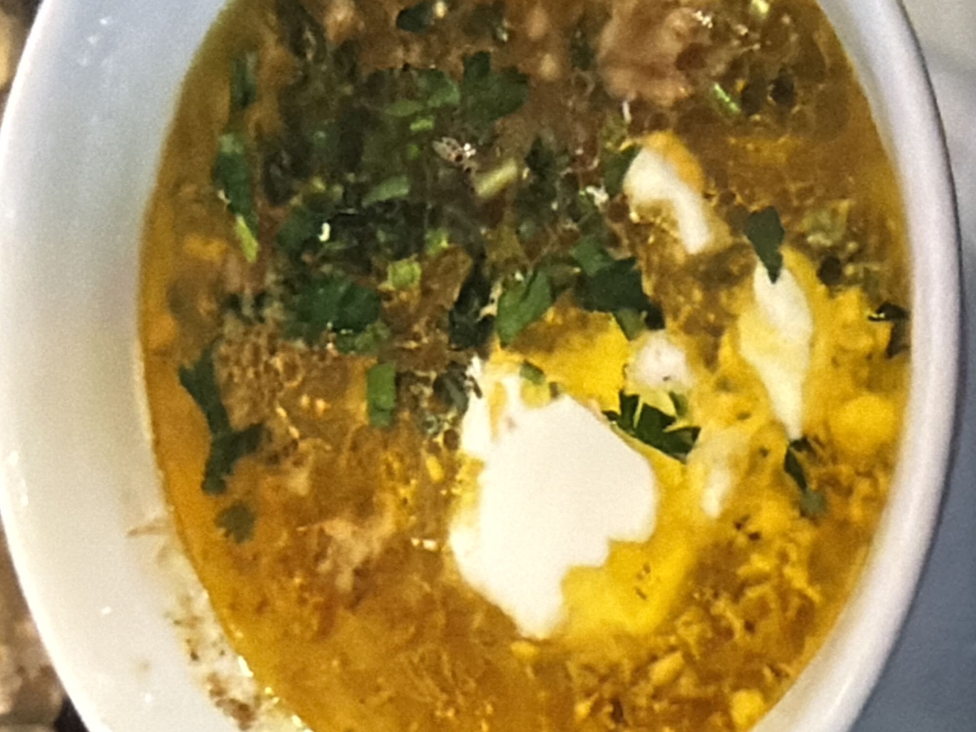 A close-up, top-down, slightly angled shot of a white bowl filled with a steaming, golden-yellow soup. The soup appears to be hearty, with visible pieces of ingredients and flecks of green herbs, likely cilantro, scattered throughout. A dollop of white, creamy substance, possibly yogurt or sour cream, sits prominently in the center of the bowl, swirling slightly into the broth. The broth has an oily sheen, indicating richness. The lighting is soft, suggesting an indoor setting, possibly a kitchen or dining area. The image is somewhat blurry, which can give it an artistic or home-style feel. No text is visible in the image. The overall impression is of a warm, comforting, and flavorful dish.