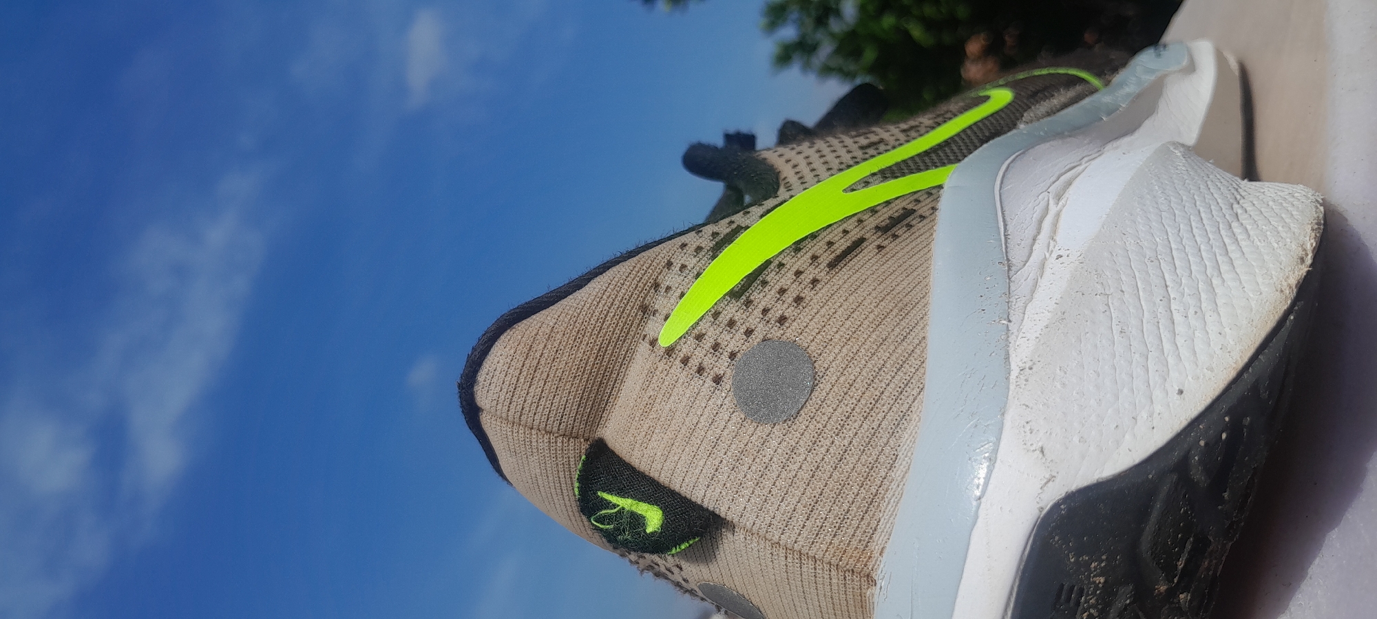 A close-up, low-angle outdoor shot features the heel and side of a tan and white athletic shoe against a bright blue sky with scattered white clouds. The shoe has a neon green Nike swoosh logo prominently displayed on its side, and a smaller neon green logo on the heel tab. A reflective grey circular patch is visible on the tan fabric. The shoe appears to be resting on a light-colored, flat surface, possibly a table or ledge. The background shows blurred green foliage and trees, suggesting an outdoor setting in a natural or park-like environment. The image does not depict any specific landmarks or recognizable features of Abuja, Nigeria, beyond it being an outdoor scene.