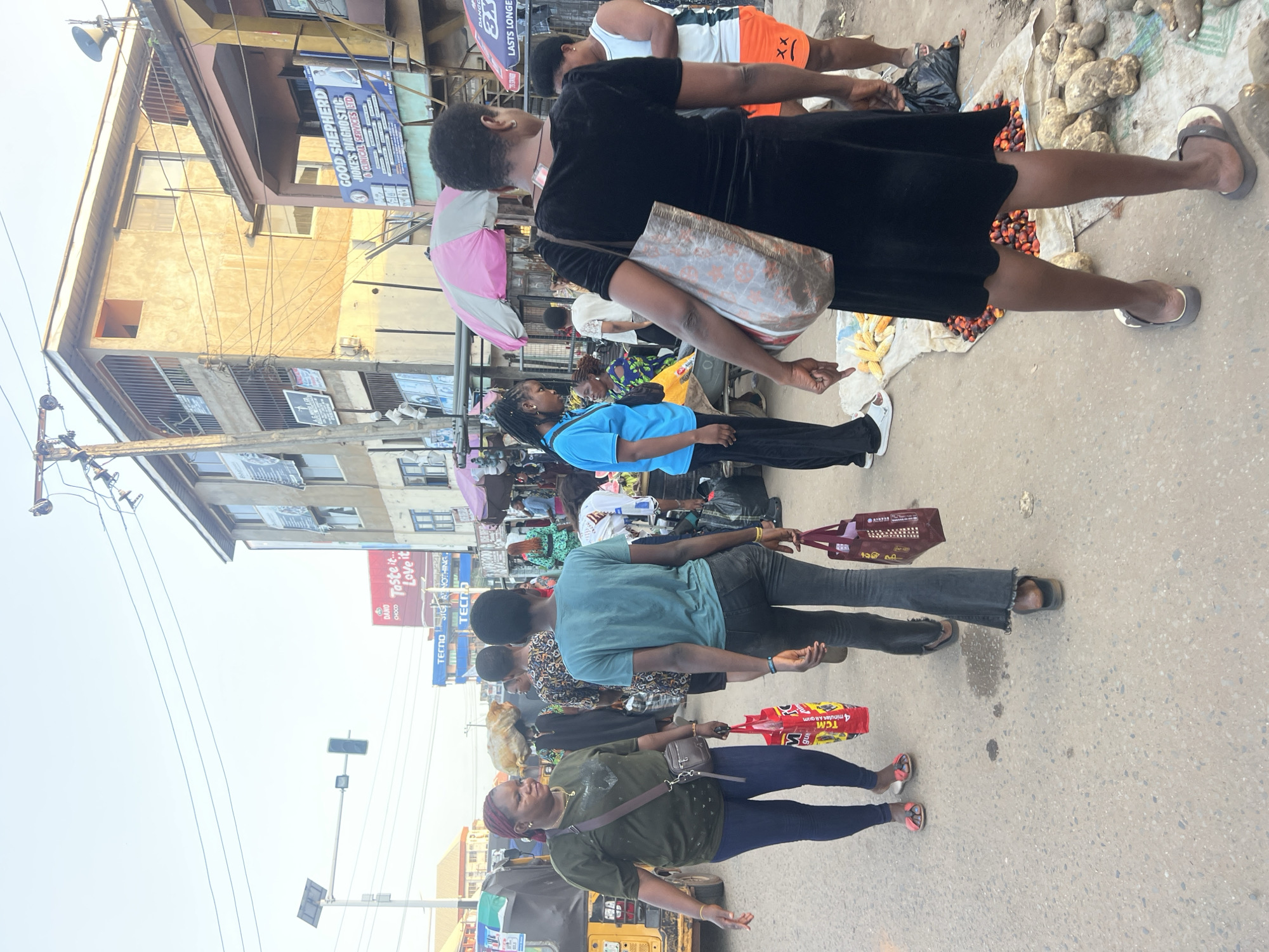The image depicts a street scene in Nkpor, Nigeria, under a hazy sky. A busy market atmosphere is evident, with several individuals navigating the paved street. In the foreground, a woman in a green t-shirt and blue jeans walks towards the left, carrying a red shopping bag. To her right, another woman in a teal t-shirt and black jeans walks in the same direction, also holding a shopping bag. Behind them, a woman in a black dress with a patterned bag slung over her shoulder is the most prominent figure. She is walking away from the camera, towards the right side of the image.
Various market stalls are visible along the street, indicated by umbrellas and displays of produce. To the left, a yellow motorized tricycle, often used for public transport, is partially visible. Buildings line the street, some appearing multi-storied with balconies and advertising posters. One building features a prominent blue and white sign for "GOOD SHEPHERD JOKES' DIAGNOSTIC & CLINICAL SERVICES LTD". Another sign reads "DANGOTE SUGAR". Power lines are strung across the top of the frame, and a traffic light is positioned on a pole. The ground is a worn asphalt surface with some debris and scattered produce, suggesting an active marketplace. The overall impression is one of a vibrant, everyday commercial environment in a Nigerian city.