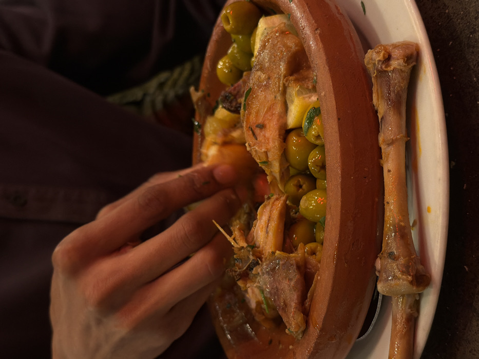 This image displays a close-up view of an individual consuming a meal from a terracotta tagine dish within a restaurant setting in Paris, France. A person's right hand, with a medium-dark skin tone, is reaching into the tagine, appearing to pick up food. The individual is wearing a dark purple or maroon long-sleeved shirt.
The tagine is filled with a stew primarily composed of pieces of chicken, numerous green olives, and slices of lemon, submerged in a rich, yellowish-orange sauce and garnished with visible herbs. Directly beneath the tagine, on a white ceramic plate, two clean chicken bones are present, indicating prior consumption of meat. Minor splatters of the orange sauce are also visible on the plate. The background is out of focus, confirming an indoor dining environment, consistent with a Moroccan or North African restaurant. The specific address within Paris is not identifiable from the visual evidence.
