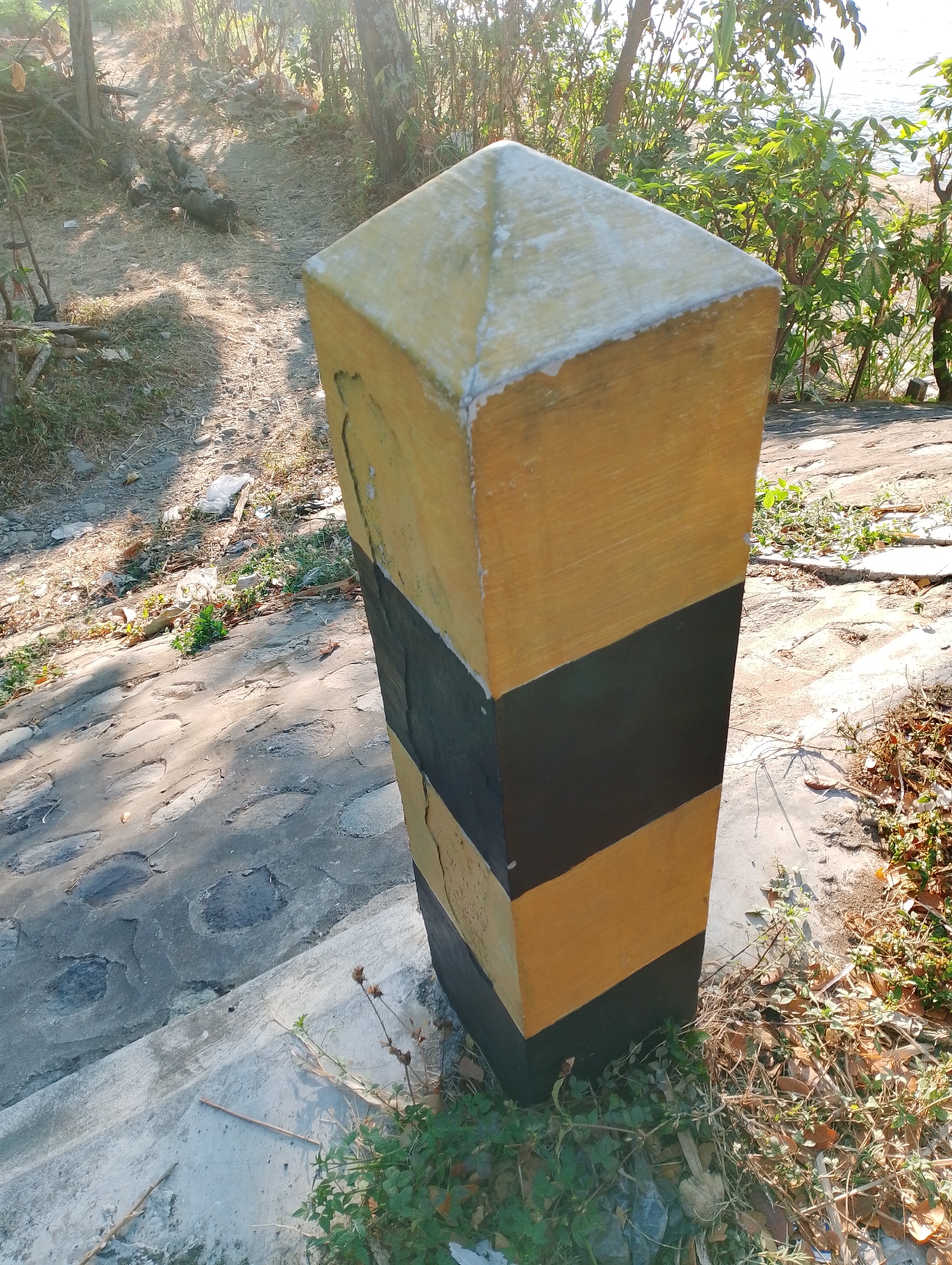 The image shows a striped marker post, painted yellow and black, standing at the edge of a paved area. The post is angled, with a pyramidal top. The paving is uneven and appears to be made of large, irregular stones. Vegetation, including dry leaves and small green plants, grows around the base of the post and along the edge of the paved area. In the background, there is a dirt path with some debris, and further beyond, lush green foliage and trees are visible, with sunlight filtering through the leaves. The scene suggests an outdoor environment, possibly in a natural or semi-natural setting near a body of water, given the hint of a shoreline in the upper right background. The location is identified as Mojo, Indonesia.