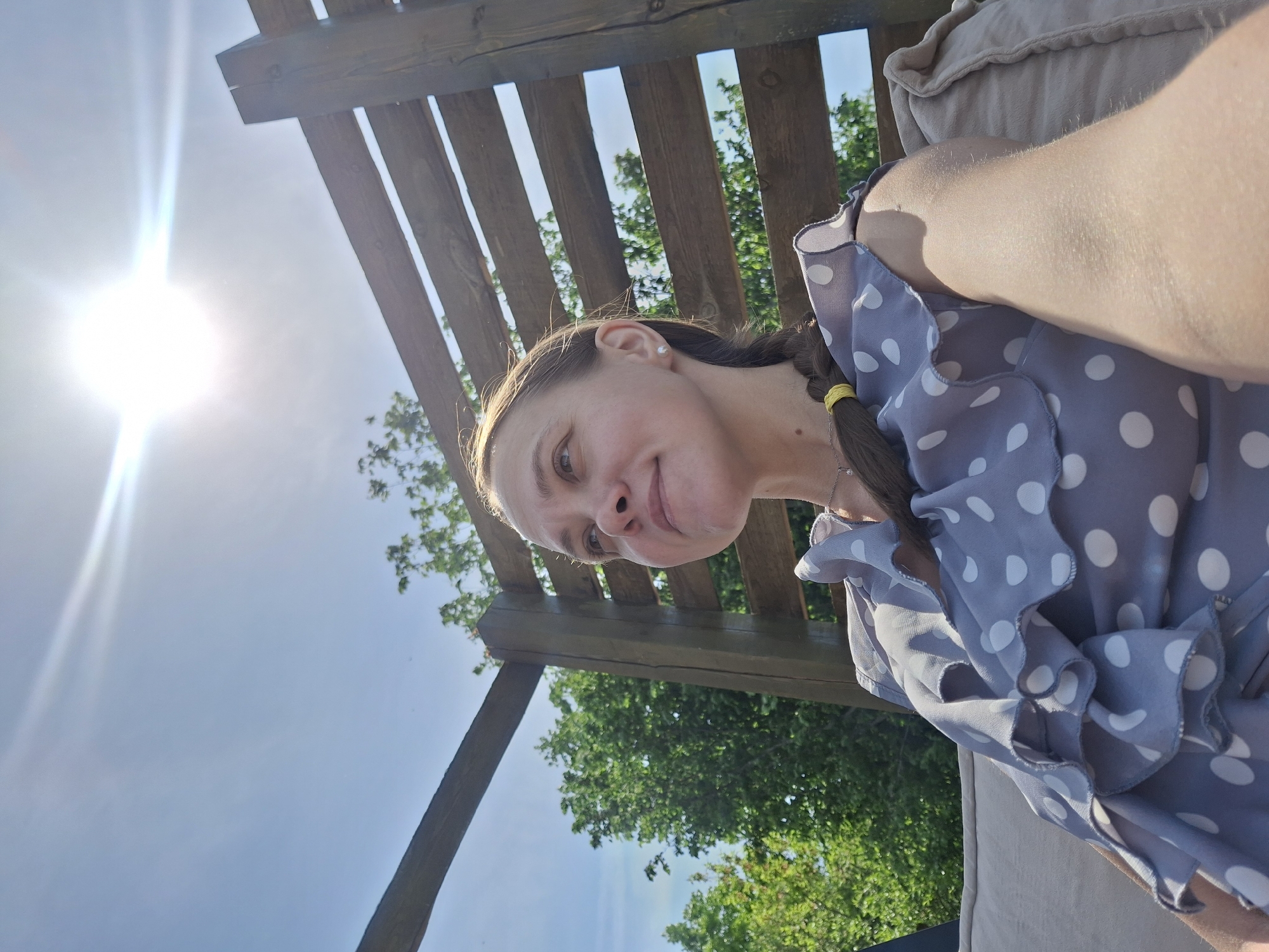 A woman is visible in the foreground, wearing a light blue polka dot shirt with ruffled edges. Her dark hair is braided and secured with a yellow tie. The background shows a wooden pergola with gaps between the slats, allowing glimpses of green trees and a blue sky. The sun is shining brightly. It's not possible to determine the exact location within the city of Chaikovsky, Russia, from this image alone.