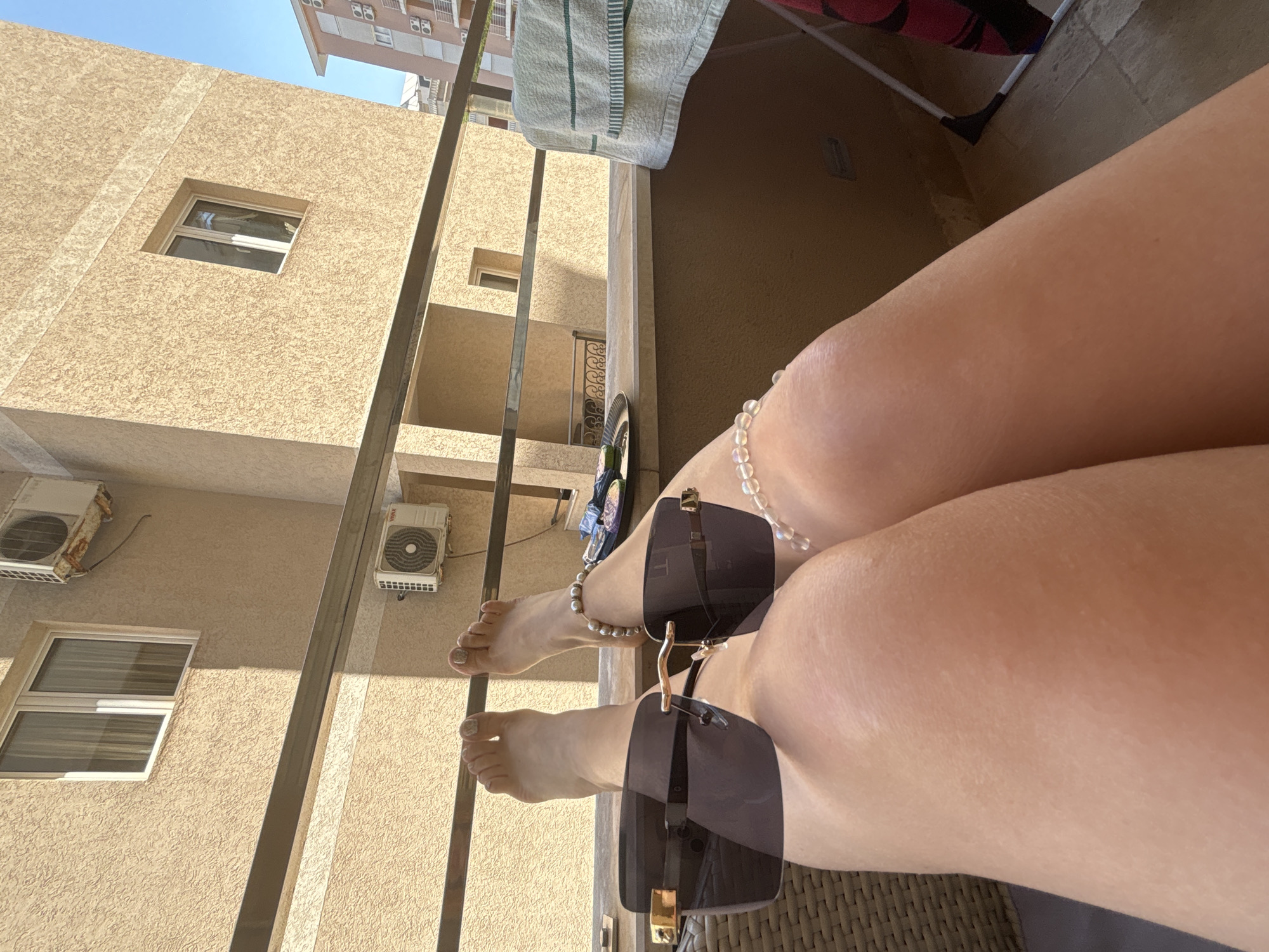 The image presents a first-person perspective from a balcony in Budva, Montenegro, during daylight hours. In the foreground, the lower legs and feet of an individual are visible, with dark-lensed, square-shaped, rimless sunglasses featuring gold accents resting on the thighs. The toenails are painted a light, possibly glittery, color, and a white beaded anklet is present on the right ankle. Part of a wicker-style chair is discernible on the left.
The mid-ground features a horizontal metal balcony railing. To the right, a metallic tray containing small, indistinct objects rests on the balcony ledge. A white towel with green stripes is draped over a partially visible white drying rack on the far right.
The background consists of several multi-story buildings with light brown, textured facades under a clear blue sky. An adjacent building displays two white-framed windows and an older, rusty outdoor air conditioning unit on its upper exterior, with a newer VIVAX branded air conditioning unit visible below the balcony railing on the left. Further right, a building with a pinkish facade and multiple balconies is also visible. Strong shadows indicate direct sunlight.