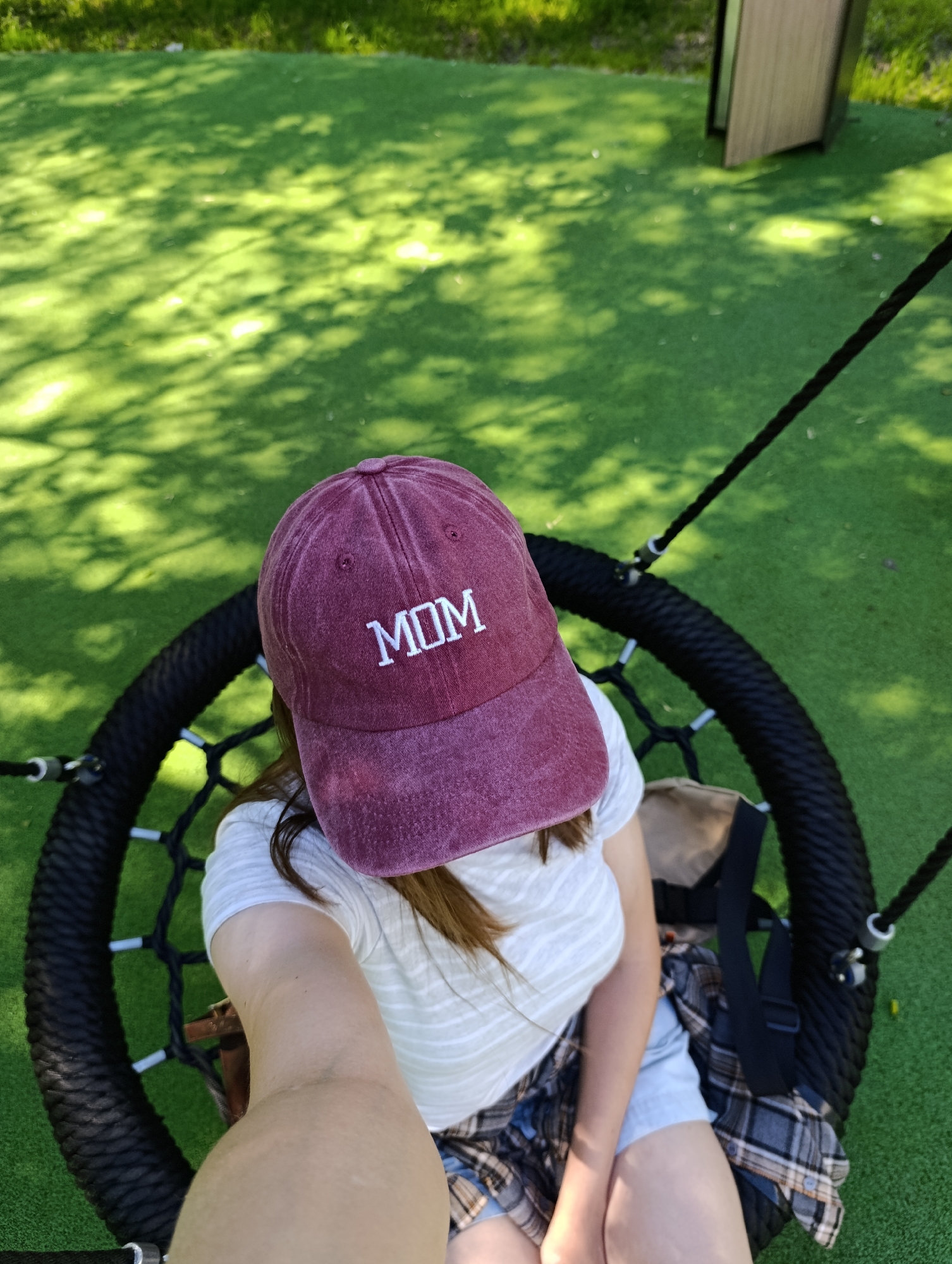 The image depicts a high-angle, self-portrait perspective of a female subject seated on a black circular nest swing in an outdoor setting in Moscow, Russia. The subject wears a faded maroon baseball cap with "MOM" embroidered in white capital letters. Below the cap, her brown hair is visible. She wears a light-colored, subtly striped t-shirt and light blue denim shorts. A plaid shirt, featuring a pattern of grey, brown, and white, is tied around her waist. A beige backpack with black straps is positioned on the swing next to her left side. The ground beneath the swing is covered in artificial green turf, displaying dappled light and shadows consistent with sunlight filtering through overhead foliage. In the upper right background, a light-toned wooden structure is partially visible. The overall scene is brightly lit by natural sunlight.