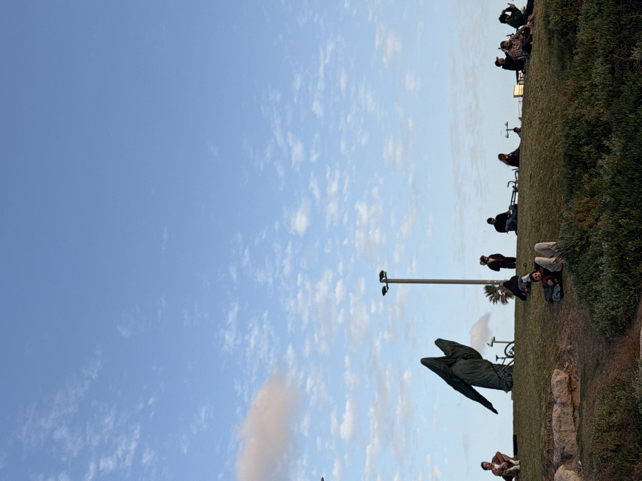 People are scattered along a grassy hill overlooking a clear, partly cloudy sky. A dark, wing-shaped sculpture stands prominently on the slope, near a light pole. Bicycles are parked nearby. Individuals sit, stand, or recline, enjoying the outdoor space. The scene suggests a peaceful gathering in a public park.