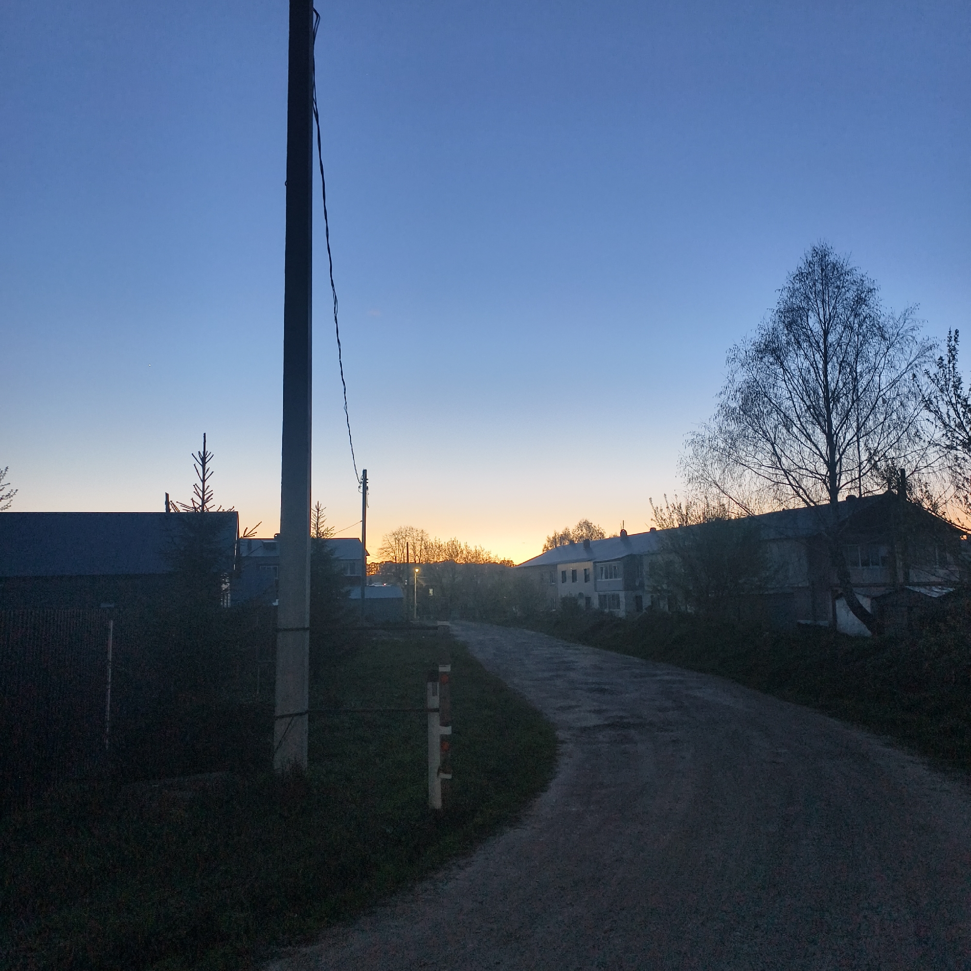 The image depicts a road in Фоки, Russia, at dusk. A utility pole stands on the left side of the road, and a safety marker is visible further down the road on the same side. Buildings line the horizon, silhouetted against the setting sun. A bare tree stands to the right of the road. The sky is a gradient of blue, transitioning to orange near the horizon. The road surface appears to be asphalt. Vegetation is visible on either side of the road. No people are visible.