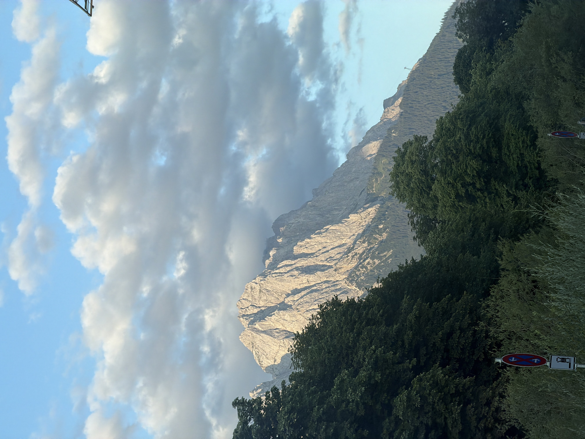 The image captures a majestic mountain range bathed in the warm glow of sunlight, likely during sunrise or sunset, due to the soft lighting highlighting the rugged peaks. The mountains are characterized by sharp, rocky formations and are partially covered with dense evergreen forests on their lower slopes. In the foreground, a lush canopy of dark green trees and dense undergrowth creates a natural framing for the scene. A traffic sign, featuring red and blue circles with arrows and a prohibition symbol, is visible on the left side, indicating a no parking or restricted area. The sky above is a serene blue, dotted with scattered clouds that are tinged with gray and white, suggesting a dynamic weather pattern. The overall atmosphere is one of natural grandeur and tranquility. This scene is highly likely to be the Zugspitze massif, the highest peak in Germany, located in the vicinity of Garmisch-Partenkirchen.