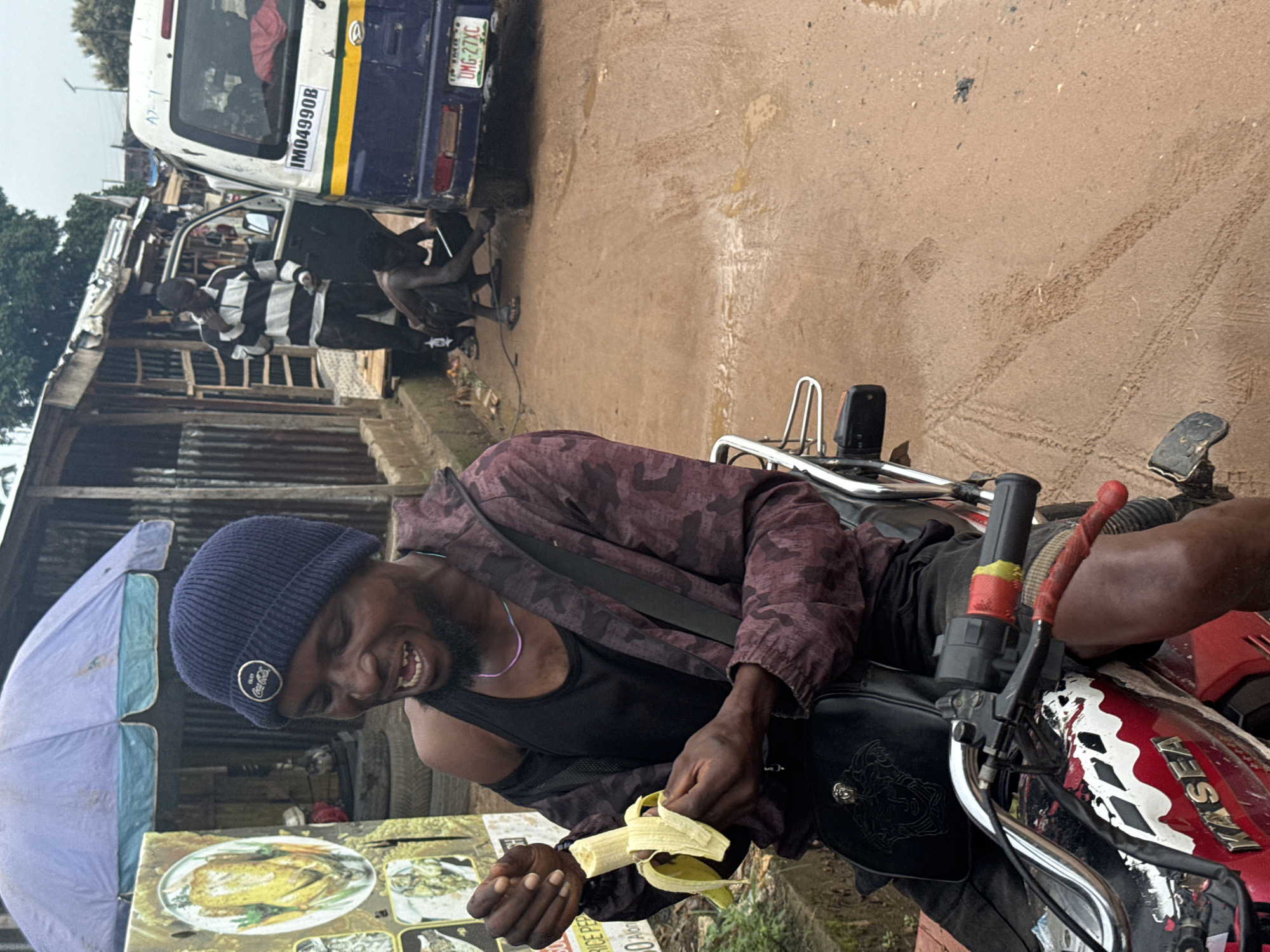 In Ihiagwa, Nigeria, a man wearing a dark blue beanie and a maroon and black camouflage jacket sits on a red and white motorcycle, peeling a banana. He wears a black tank top underneath his jacket and a thin, iridescent necklace. A black bag with a gold emblem is slung across his chest. The motorcycle has the word "KASEA" in white lettering on its side. In the background, a group of people interact near a blue and white van with license plate IM04990B and a sticker reading UMG-27XC. One man in a black and white striped shirt stands with his hand near his face, while another crouches down to work on the van's rear tire. The environment appears to be an outdoor market or roadside stall, indicated by the corrugated metal structures and a large umbrella overhead. The ground is muddy and wet, with tire tracks visible in the dirt. A poster displaying food items is propped up to the left of the man on the motorcycle. The sky is overcast.