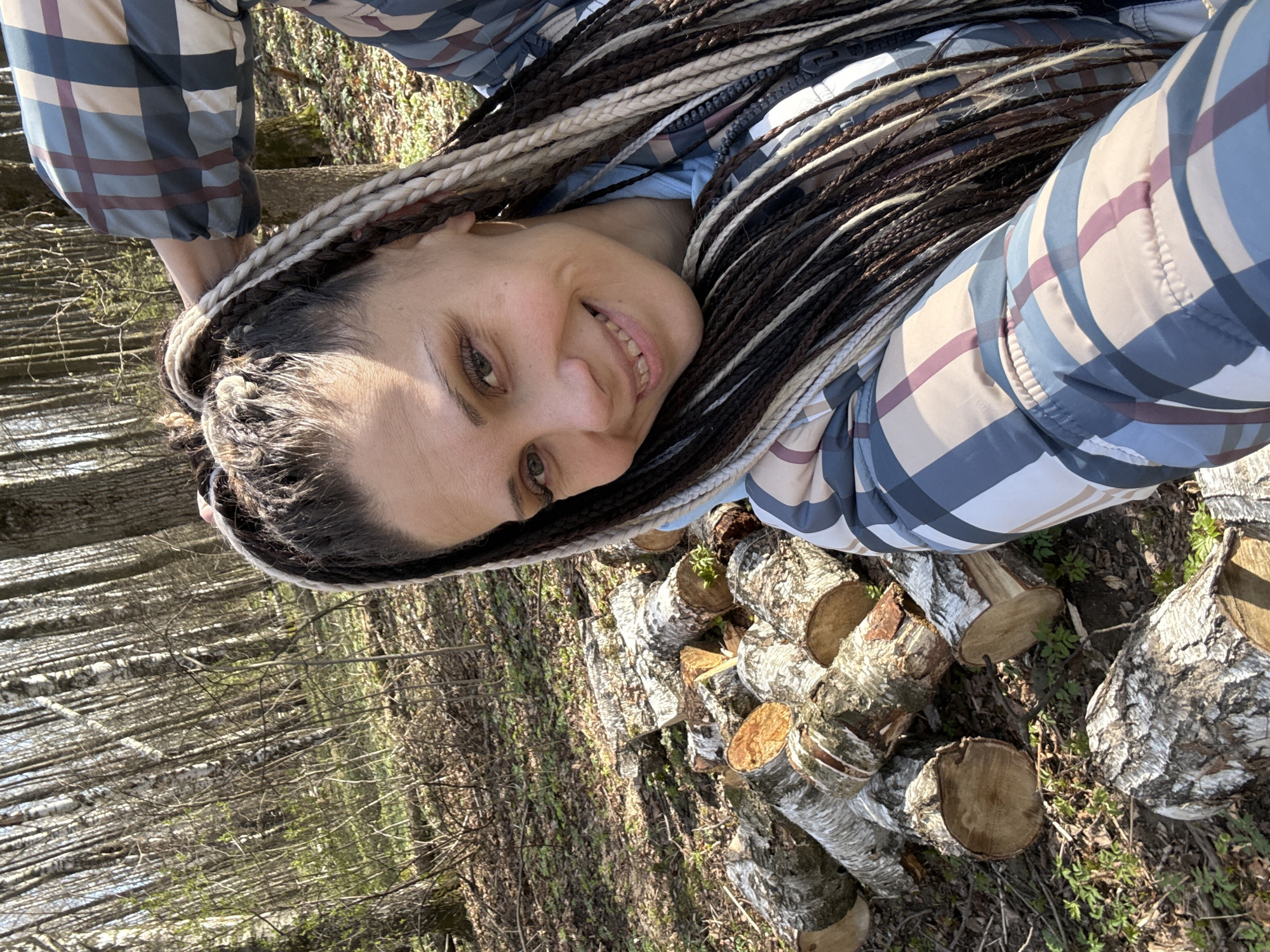 A medium close-up, eye-level selfie captures a woman smiling broadly at the camera, her head tilted to her right. She has long, dark and light-colored braided hair (dreadlocks or similar) that frames her face and extends over her shoulder. Her light-colored eyes are open and engaging. She is dressed in a plaid jacket featuring blue, beige, and maroon squares, worn over a light blue top.
The setting is an outdoor, wooded area, likely a forest, under bright daylight. In the foreground and midground, a pile of freshly cut logs, identifiable as birch by their distinct bark, are scattered on the ground. The ground cover includes fallen dry leaves, some patches of small green plants, and dry grass. The background reveals numerous deciduous trees with bare branches, suggesting the season is either late winter or early spring. The scene conveys a sense of outdoor enjoyment in a natural environment. No visible text is present in the image. The location context provided is Rusanovo, Russia.
