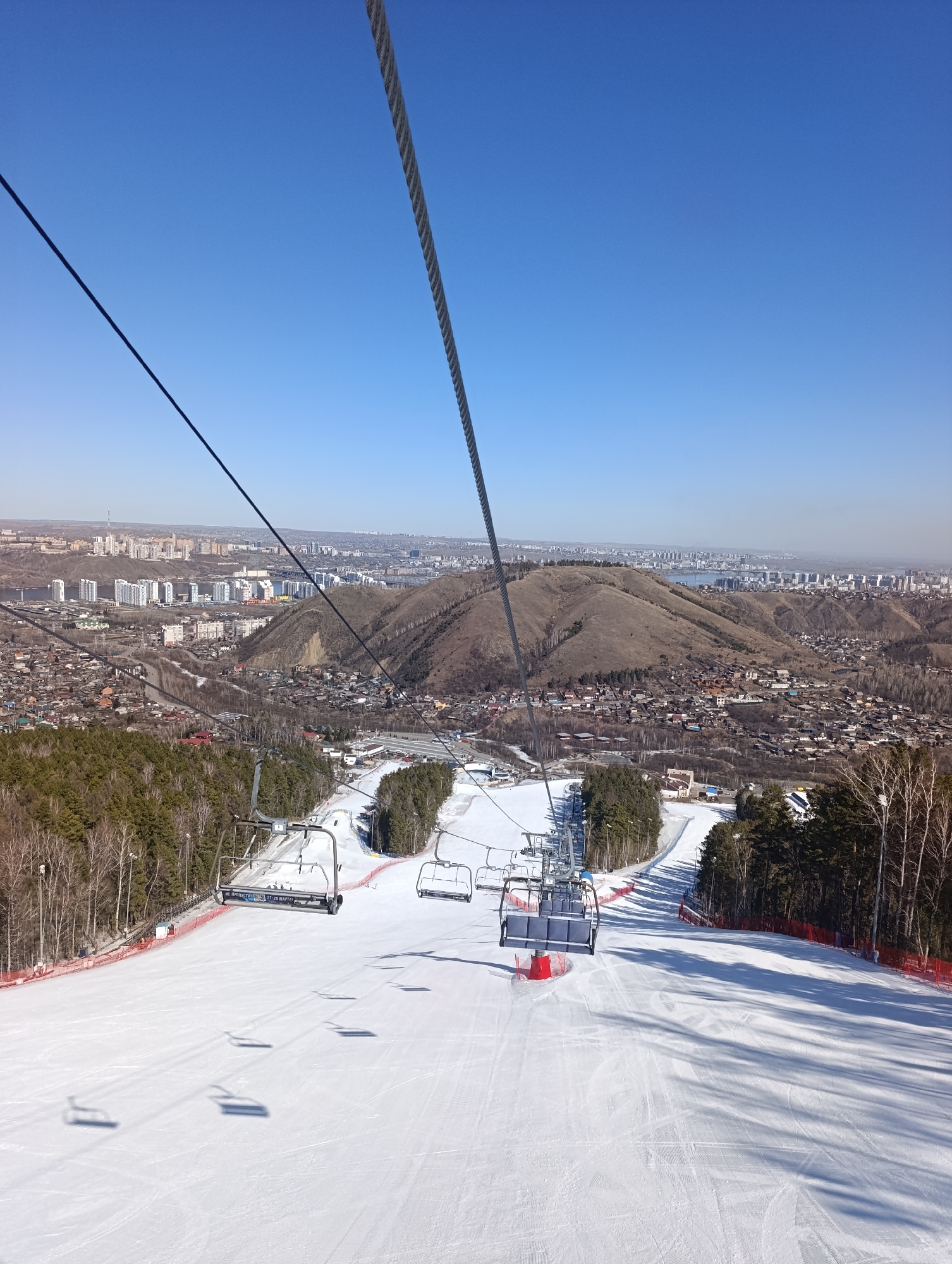 The image depicts a ski slope from the perspective of a ski lift. The foreground is dominated by a snow-covered slope with visible tracks. Several ski lift chairs are suspended above the slope. In the midground, a landscape of buildings, trees, and hills stretches out. The cityscape in the distance suggests the location is likely a city with mountainous or hilly terrain. The sky is clear and blue, indicating a sunny day. Based on the style of the buildings and the overall landscape, it is likely that the picture was taken from Bobrovy Log in the city of Krasnoyarsk, Russia.