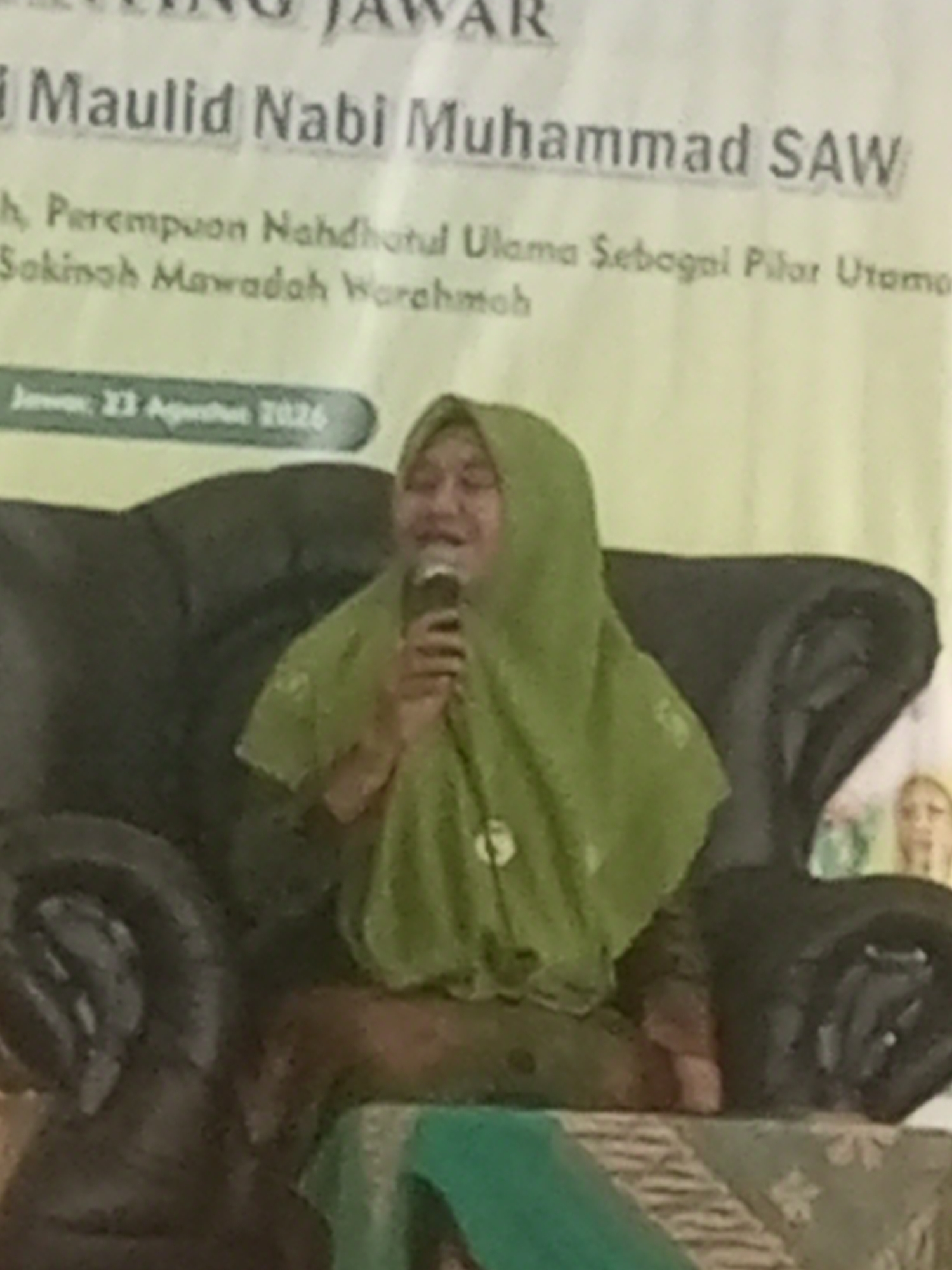 The image shows a woman wearing a green hijab and a matching green garment, seated on a dark-colored sofa. She is holding a microphone to her mouth, as if speaking or singing. Behind her, a banner is visible with text that appears to be in Indonesian, including "Maulid Nabi Muhammad SAW" and "Perempuan Nahdlatul Ulama Sebagai Pilar Utama Sakinah Mawaddah Warahmah". Below this, there is text that reads "Jawa, 23 Agustus 2026". The background is a light green wall, and on the right side, a partial image of another person's face is visible, wearing a light-colored garment. The overall scene suggests an event or presentation taking place. The event appears to be held in Klaseman, Indonesia, as indicated by the text on the banner.