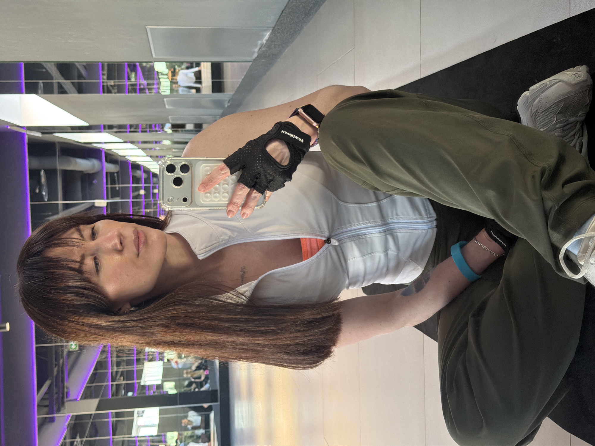 A woman takes a selfie in a gym. She is wearing exercise gloves, athletic pants, and a white vest. The background shows gym equipment in a mirrored wall.