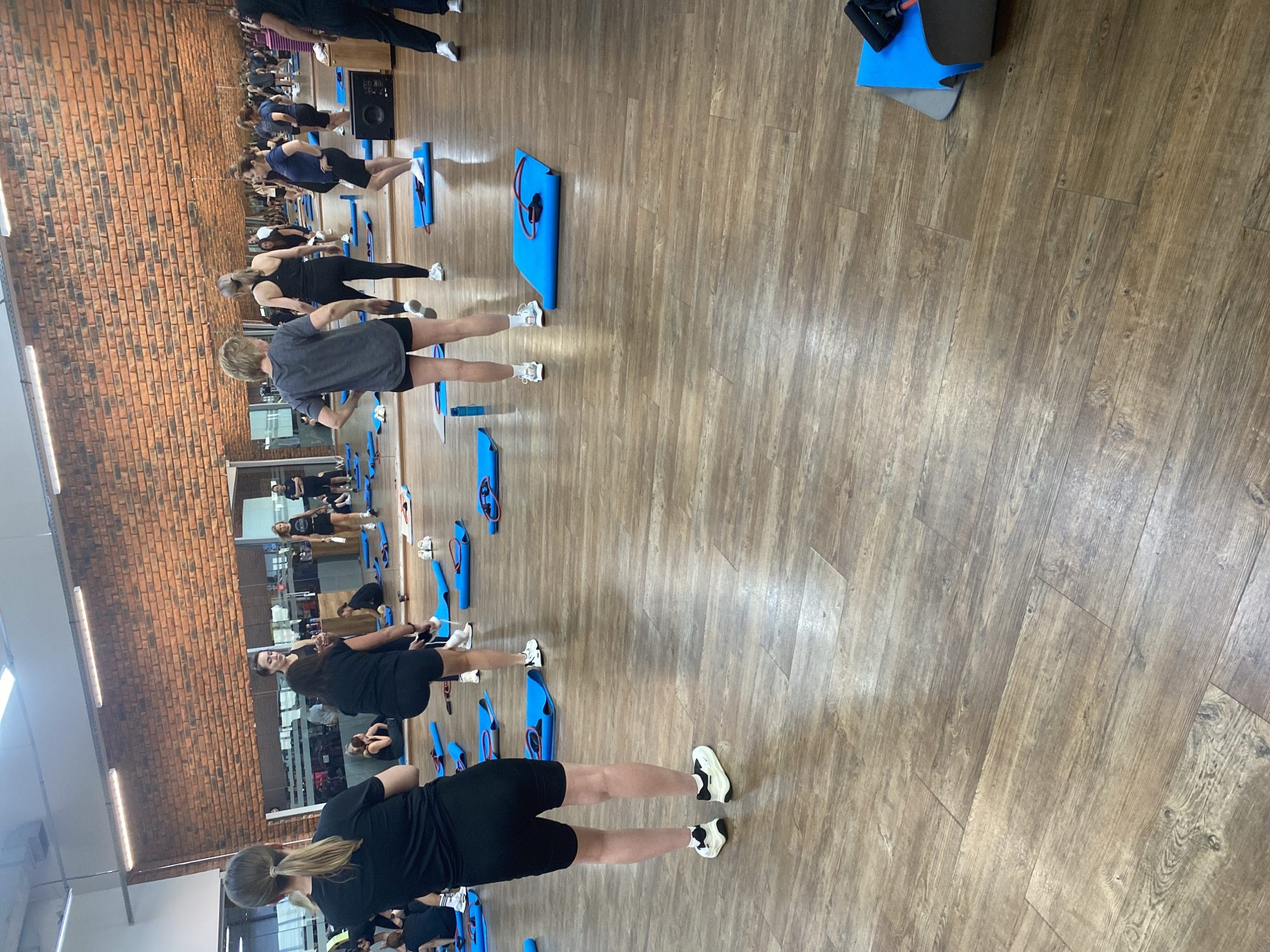 This is a wide shot of a fitness class in progress in an indoor gym. The floor is polished wood. The back wall is a brick wall. On the left side, a woman with her back to the camera, wearing a black t-shirt and black shorts, stands facing a group of women and a female instructor. Several other women are scattered throughout the room, some on blue exercise mats with resistance bands. The room is well-lit with fluorescent lighting fixtures on the ceiling. A large mirror runs along the back wall, reflecting the scene.