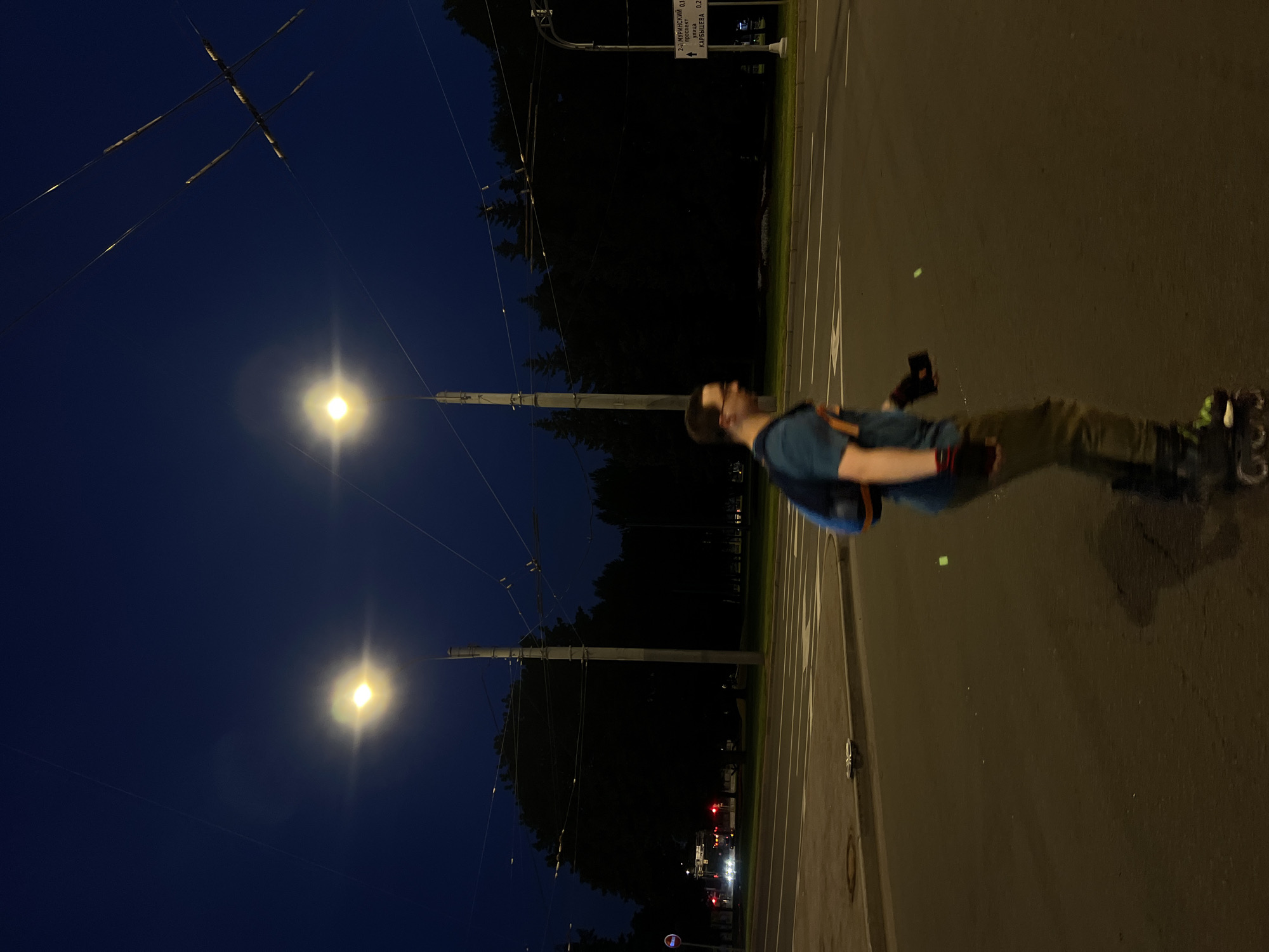 The image depicts a nighttime scene in Saint Petersburg, Russia. A person is rollerblading on a street at dusk, with streetlights illuminating the scene. The person is wearing a blue t-shirt, dark pants, and rollerblades. They are also carrying a backpack. The street is a multi-lane road with traffic lights and overhead trolleybus wires visible. In the background, there are trees and buildings, suggesting an urban environment. A sign in Russian, likely a street name or direction, is visible on the right side of the image. The sky is dark blue, indicating nighttime.