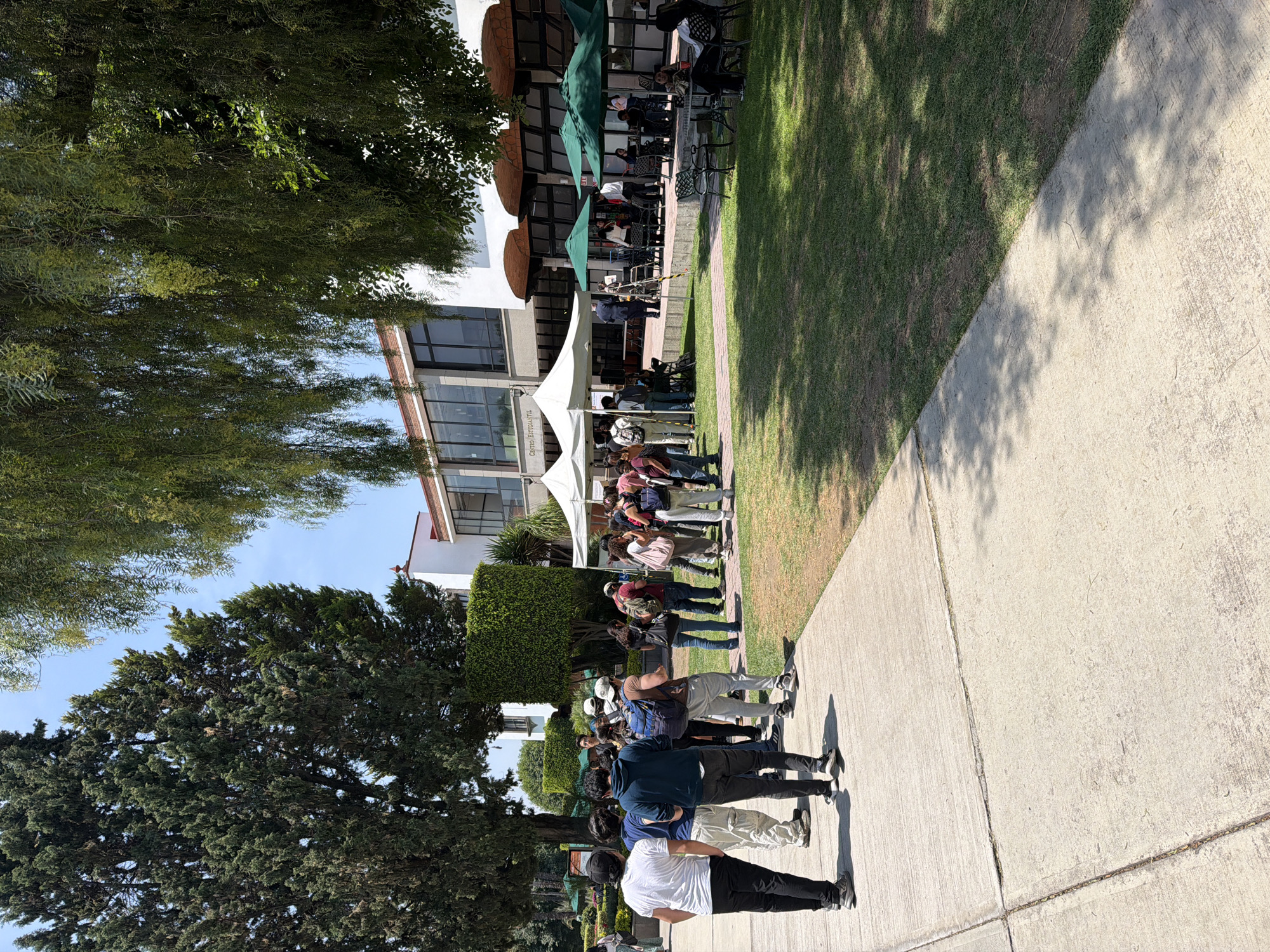 This outdoor scene, set in San Andrés Cholula, Mexico, captures a lively gathering on a sunny day. A crowd of people, predominantly young adults, are seen congregating and moving along a concrete pathway that runs parallel to a grassy area. Many individuals appear to be standing in lines or groups, suggesting an organized event.
To the left, a modern, multi-story building with white walls and dark-framed windows forms the backdrop. Its exterior features areas with shaded outdoor seating, complete with tables and green umbrellas, where some people are relaxing. Near the front of the building, a large white tent is set up, under which more people are gathered. Visible on the side of this tent is the text "Centro Literaril," indicating a literary or cultural event.
The surroundings are lush with greenery. Dense, dark green trees line the left side of the frame and appear further in the background, contrasting with the bright blue sky. Manicured hedges also contribute to the park-like or campus ambiance. The sun casts long, distinct shadows across the concrete path and the grass, suggesting it is either morning or late afternoon with clear weather. The ground transitions from a well-maintained lawn on the right to the light grey concrete path where most of the people are located. The overall atmosphere is relaxed and engaging, typical of a community or educational outdoor event.