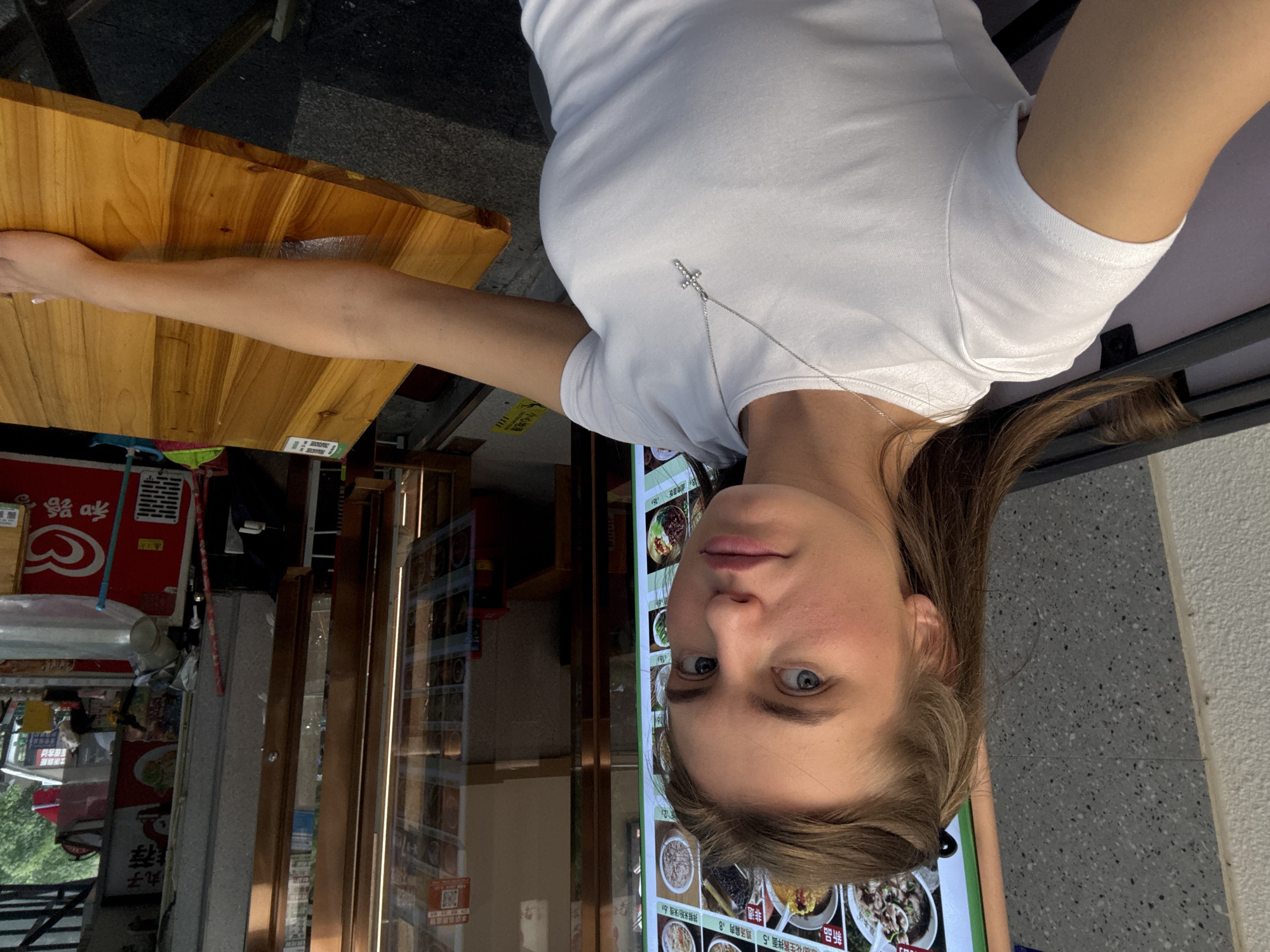 A young woman with blonde hair tied back is captured in a medium close-up shot, seated at a wooden table outside what appears to be a food establishment. She is wearing a white t-shirt and a silver cross necklace. Her gaze is directed towards the camera, and her expression is neutral. To her left, a large digital menu board displays various food items. In the background, through a glass door, a glimpse of the restaurant's interior is visible. Further to the right, a red refrigerator with an ice cream logo is present. The overall setting suggests an outdoor seating area of a restaurant or food stall in a commercial area. The location is identified as Haizhu District, China.