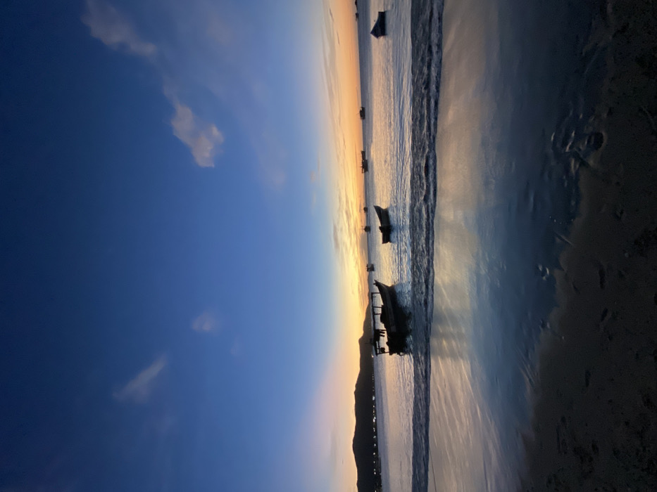 A serene beach scene is captured during a sunset. A line of silhouetted boats rests near the water's edge as waves gently lap the shore. The sand reflects the warm hues of the setting sun. The sky transitions from a deep blue to an orange glow, with scattered clouds enhancing the peaceful atmosphere. A dark, silhouetted mountain is visible in the background.