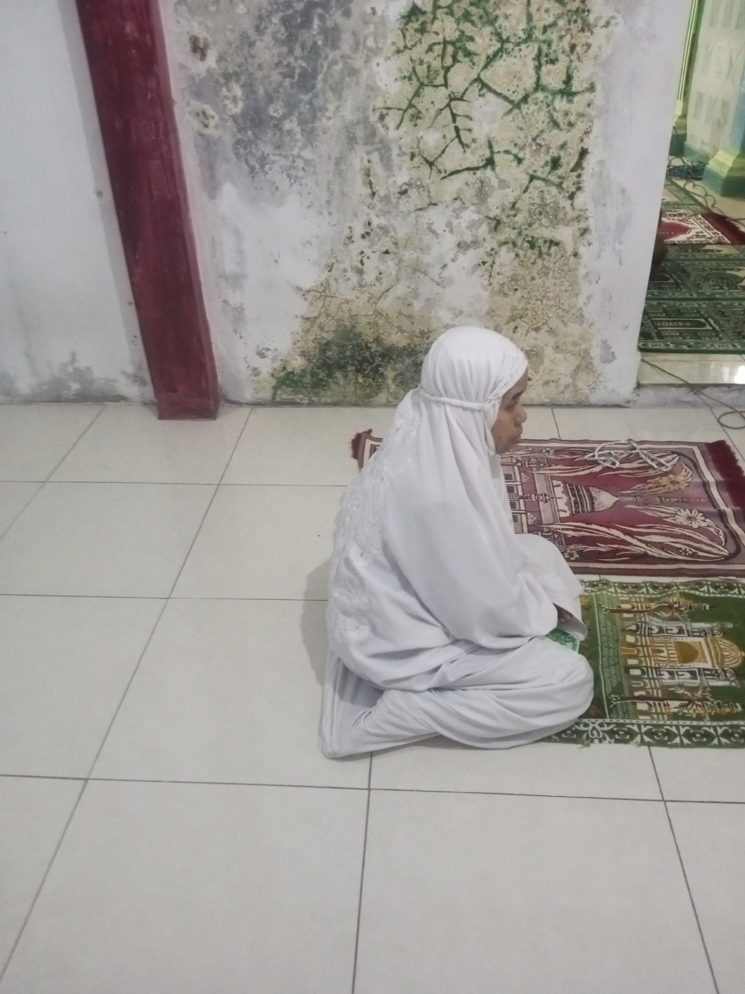 A woman wearing a white hijab and a white prayer dress is sitting on the floor in a room. She is facing right and appears to be praying. She is on a prayer mat, and there is another prayer mat to her right. The floor is tiled and white. The wall behind her is old and stained with green mold. To the left of the wall, there is a red vertical beam. The room appears to be indoors, possibly a prayer room or a home. The image does not contain explicit information to determine the specific location in Andongsili, Indonesia.