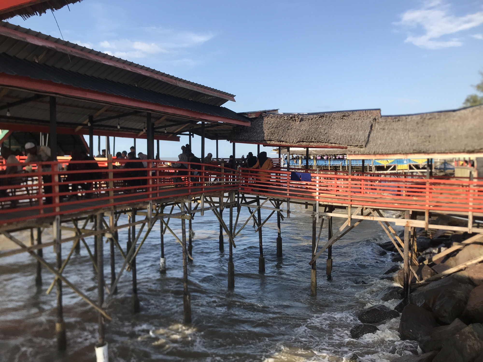 The image shows an elevated pier or platform structure extending over water. The structure appears to be a restaurant or gathering place, as people are visible on the platform. The structure is made of wood, with red railings and a combination of thatched and corrugated metal roofing. The supports for the pier are also wooden and are submerged in the water. Waves are visible, indicating a coastal location. Rocks line the edge of the platform where the pier meets the land. The image is taken in Lancok, Indonesia.