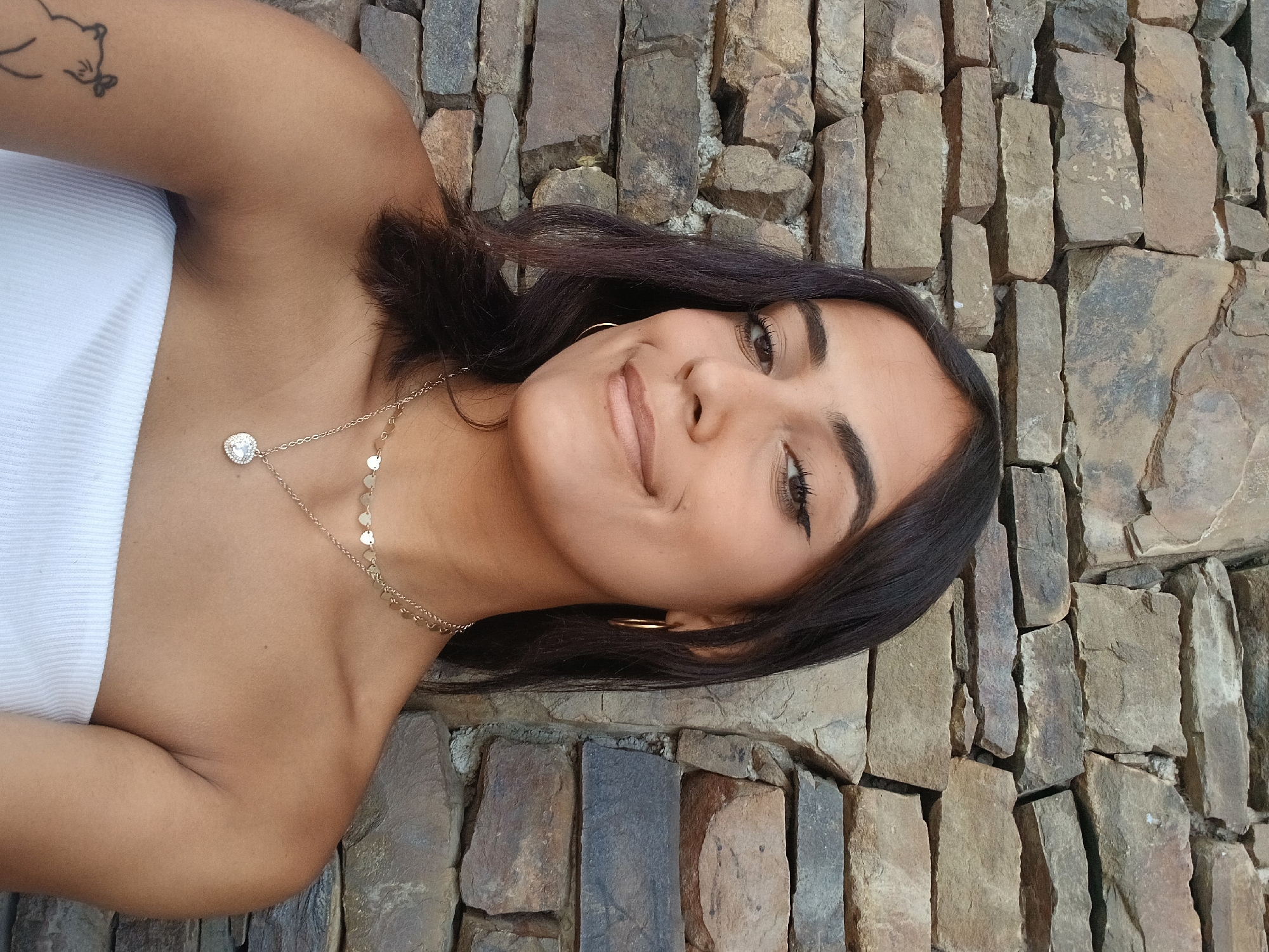 The image is a portrait of a woman with dark hair, wearing a white strapless top and a gold necklace. She is smiling slightly. The background consists of a wall made of uneven, variously colored stones. While the image provides no specific visual clues to pinpoint a location in Juan Griego, Venezuela, the stone wall could potentially be found in various settings, such as a building facade, a garden wall, or a retaining wall in a public area. Without additional visual information, determining the precise location within Juan Griego is impossible.