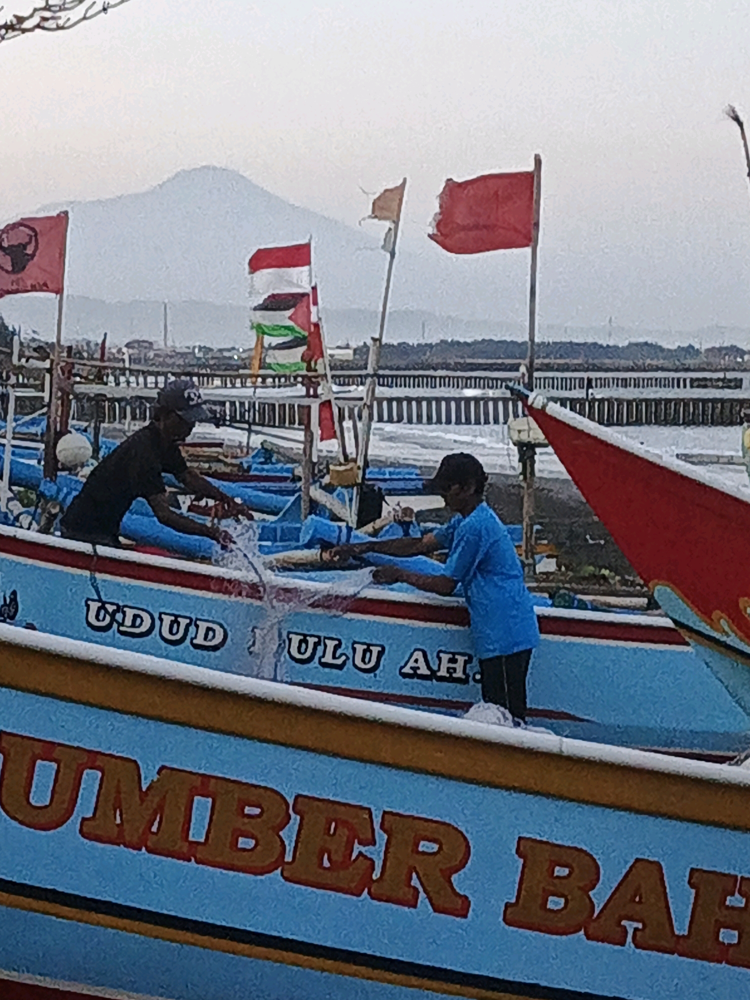 The image depicts a group of fishing boats docked at the Kelurahan Cilacap, Kecamatan Cilacap, Indonesia. In the foreground, two individuals, appearing to be fishermen, are engaged in work on one of the boats. One man, wearing a dark t-shirt and a cap, is leaning over the side of a light blue boat with red trim, seemingly handling a net. Another person, in a bright blue shirt, stands further back on the same boat. The boat is adorned with lettering, partially visible as "UDUD ULU AH" and "UMBER BAH". Behind the boats, a long, dark pier or walkway stretches across the water, with a hazy cityscape and a prominent mountain in the background. Several flags are also visible, including red flags, one with a black bull emblem, and a multi-colored flag that appears to be the Palestinian flag. The overall scene suggests a working harbor environment.
