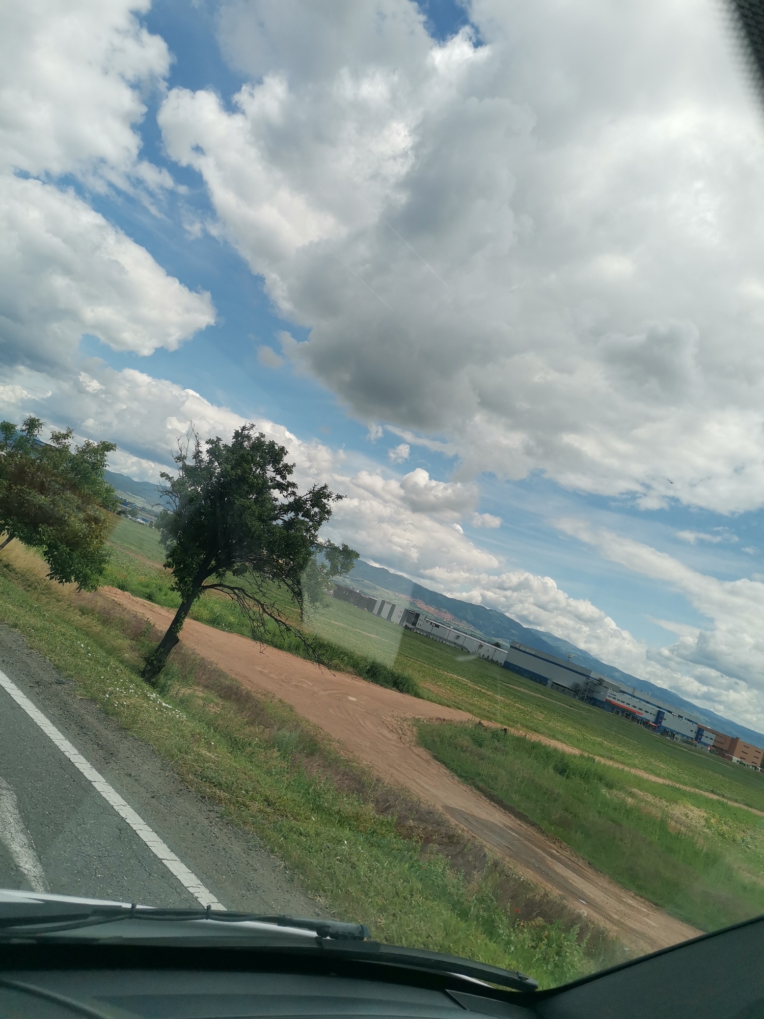 This is an outdoor, eye-level shot taken from inside a vehicle. The scene depicts a rural landscape with a road in the foreground, a field, and some buildings in the distance. The sky is partly cloudy with a mix of white and dark clouds. The road is asphalt with a white line marking the lane. There are two trees to the left of the road, one with sparse leaves and the other with denser foliage. Beyond the trees, a field stretches out, with patches of green grass and brown dirt. In the background, a row of industrial buildings with white and blue facades can be seen, along with some smaller, colorful structures. Mountains are visible in the far distance. The image appears to be taken during the daytime under natural light. It is highly probable that this image was taken on the outskirts of Sofia, Bulgaria, given the common landscape composition of industrial zones bordering fields and mountains.