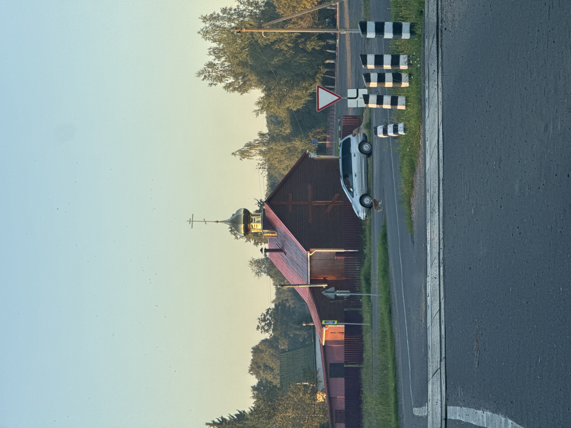 A wide shot captures a rural scene in Ходасаўскі сельскі Савет, Belarus, under a clear, pale blue sky that transitions to a soft yellow near the horizon, suggesting early morning or late evening light. In the mid-ground, a wooden building with a vibrant red roof and a golden dome topped with a cross stands out. This structure appears to be a church or religious building. To the left of the building is a dark red fence.
In front of the building, a silver minivan is parked on the paved road. A small, light-brown dog is visible near the front of the minivan, facing away from the camera. A yield sign, a triangular red-and-white sign, is positioned on a pole to the right of the minivan, near the road. Further to the right, a series of striped black and white bollards are lined up along the edge of the road. The road itself features a pedestrian crossing made of metal grating.
The background is filled with dense green trees, characteristic of a wooded rural landscape. The overall impression is one of tranquility and a typical village setting in the Belarusian countryside.