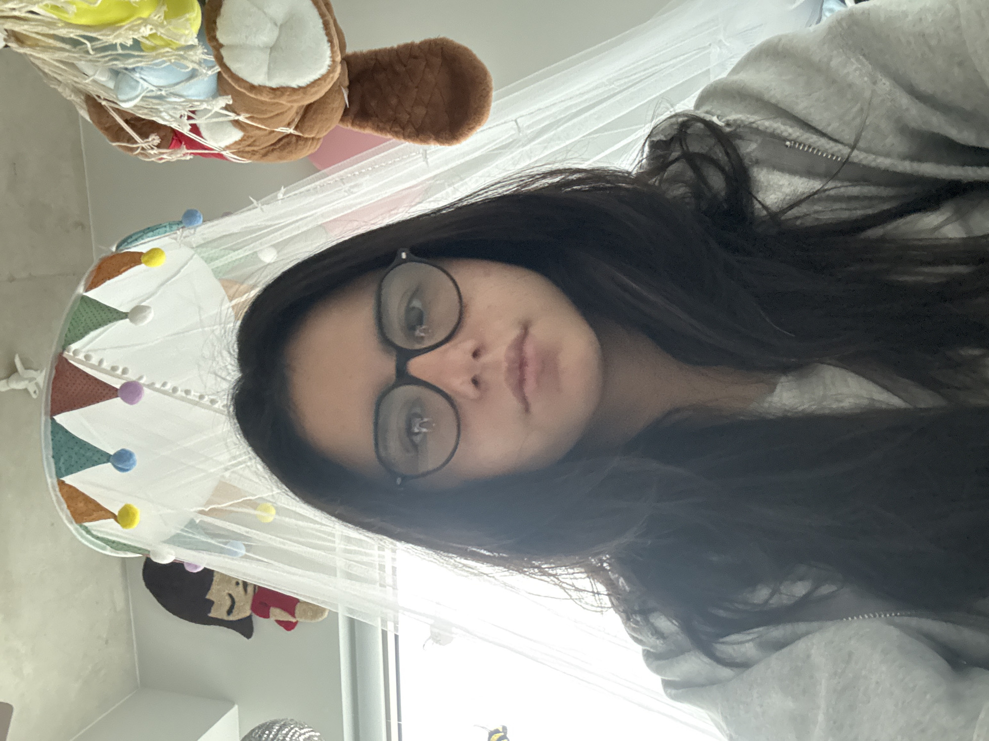A person with dark hair and glasses is looking at the camera. They are wearing a gray hooded sweatshirt. Above them, there is a child's mobile and netting, with a teddy bear and fabric shapes visible. The background suggests a bedroom or child's room. The location cannot be determined to be in Austin, United States of America.