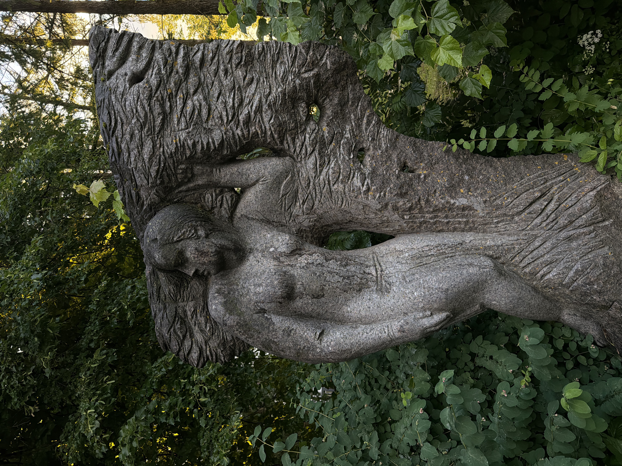 The image shows a weathered stone sculpture of a female figure supporting a rough, textured stone structure that resembles a tree trunk or a natural rock formation. The sculpture is made of grey granite and exhibits a textured, somewhat abstract style. The figure appears to be nude from the waist up, with her hair styled in waves. She is positioned with her left arm raised, supporting the stone structure, and her body is turned to the side, looking towards the left. Her lower body is partially obscured by the stone structure and the surrounding foliage. The stone itself has a coarse texture with visible striations and some yellow lichen or moss growing on it.
The sculpture is situated outdoors, surrounded by lush green foliage. Dense bushes and leafy plants frame the sculpture on the left and right sides, with some greenery extending behind it. In the background, taller trees are visible, with sunlight filtering through their leaves, suggesting it is daytime. The lighting is natural, casting some shadows and highlighting the textures of the sculpture and the surrounding vegetation. The overall impression is one of a naturalistic setting integrated with art.
This sculpture is located in the city park of Solnechnogorsk, Russia, specifically within the "Park of Heroes of the Soviet Union". It is a memorial dedicated to the victims of fascism and is known as "The Prisoner of Fascism".