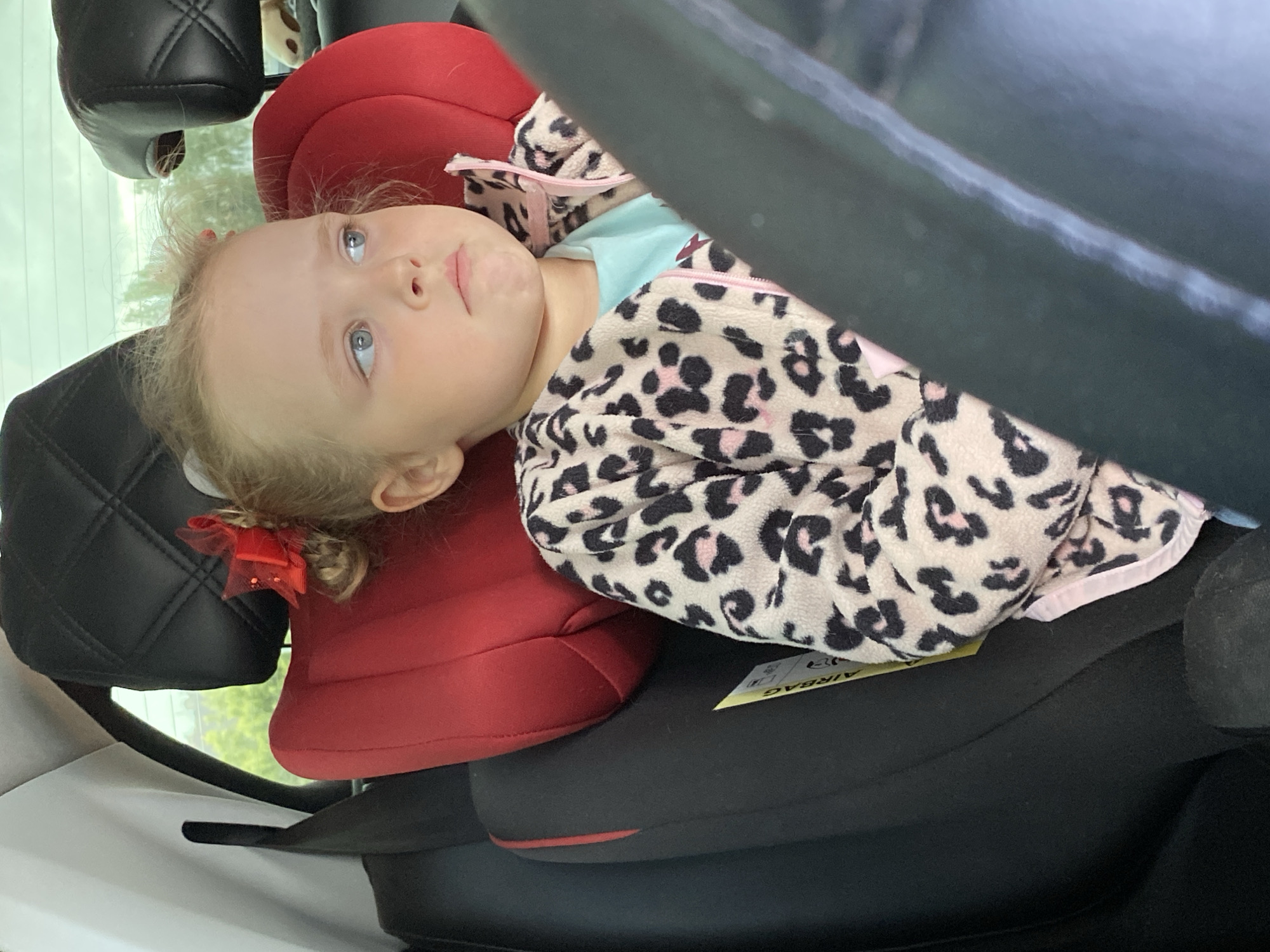 A young child, possibly a toddler, is seated in a red car seat inside a vehicle. The child has blonde hair and is wearing a leopard-print jacket over a light blue shirt. A small red bow is attached to the child's hair. The child is looking upwards with a contemplative expression. The car seat is placed on a black car seat with an "AIRBAG" tag visible. Parts of the vehicle's interior, including a black headrest and other sections of the car's interior, are visible. There is no indication of Votkinsk, Russia in the image.