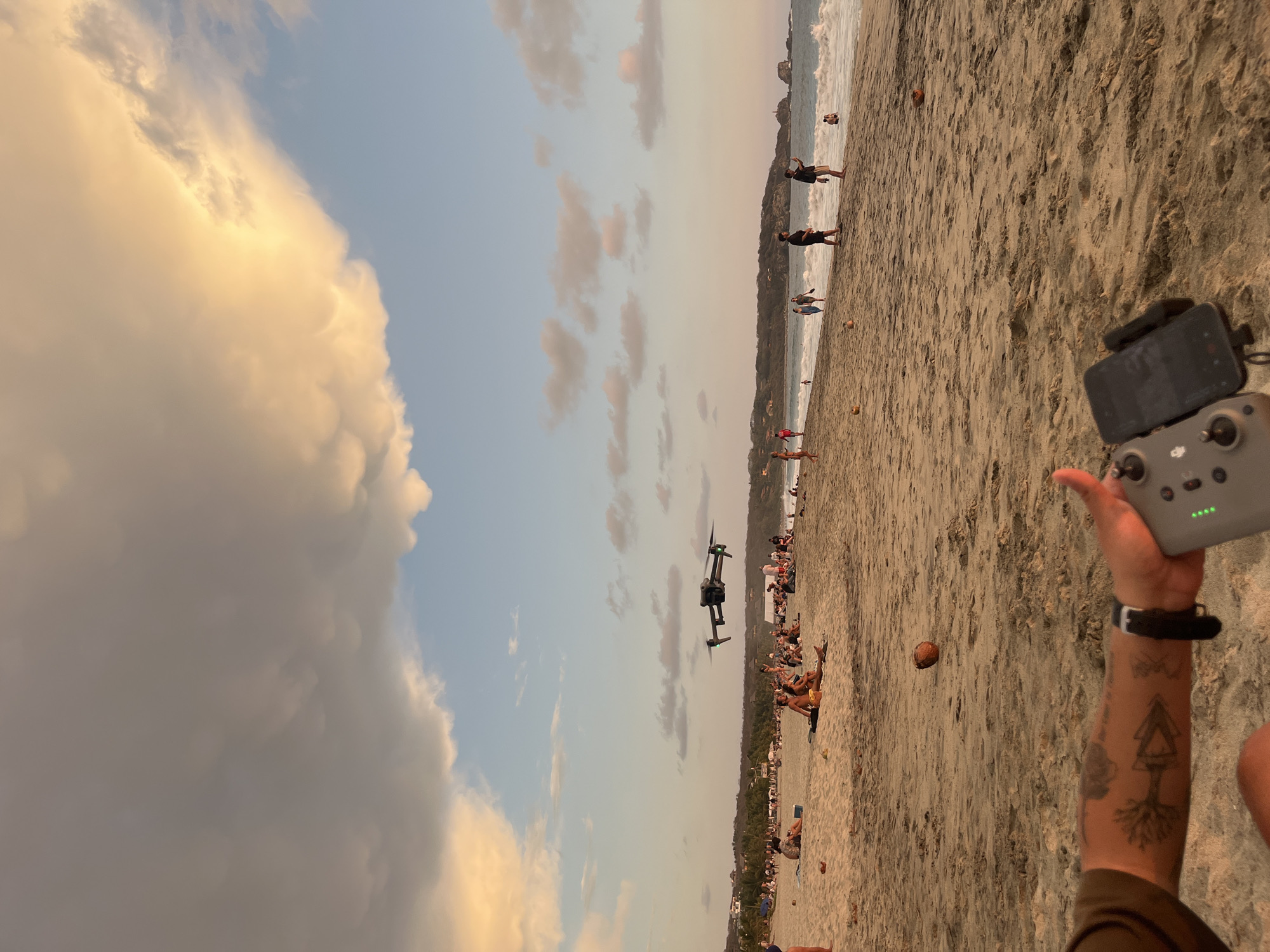 The image depicts a beach scene in Brisas de Zicatela, Mexico, during the late afternoon or early evening. The sky is partly cloudy with warm-toned clouds on the left side and lighter clouds scattered across the center.
A drone is in flight, hovering over a crowded section of the beach. People are sunbathing, walking, and engaging in various activities along the shoreline. Coconuts are scattered on the sand.
In the foreground, a person holds a drone controller attached to a smartphone. The person's arm is visible, adorned with tattoos. The sand is textured and dry near the person, transitioning to a smoother, potentially wetter surface closer to the water. Waves are visible in the background.
