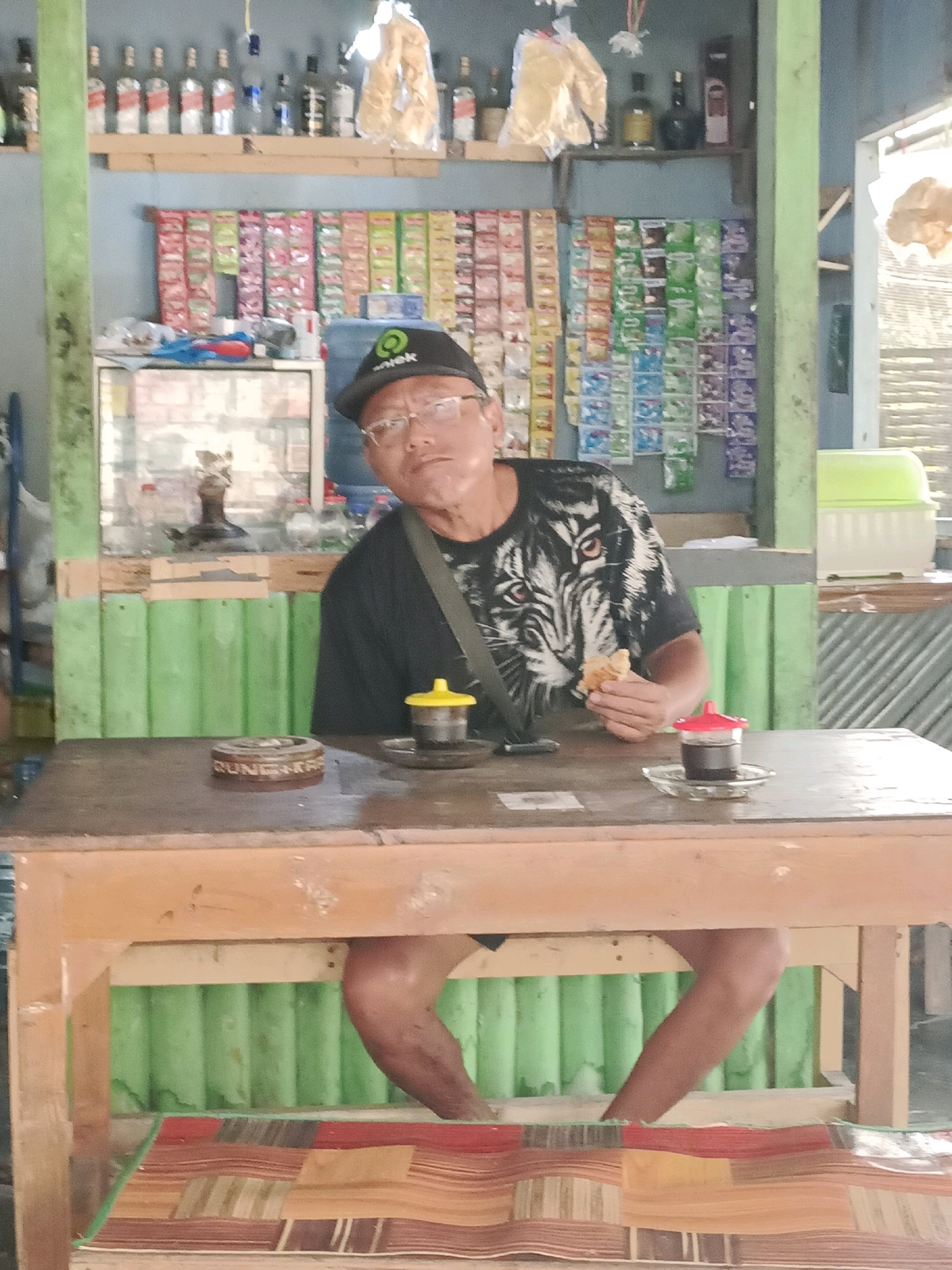 A middle-aged man wearing glasses and a black baseball cap with a green logo is seated at a wooden table in what appears to be a small shop or warung. He is wearing a black t-shirt with a white tiger graphic. He holds a piece of food in his right hand, which is near his mouth. In front of him on the table are two small cups of dark liquid, one with a yellow lid and another with a red lid, placed on saucers. An ashtray with the words "Djarum" visible is also on the table. Behind the man, shelves are stocked with various items including bottles, packaged snacks, and water jugs. The shop's walls are painted a light blue, with green corrugated metal siding visible in the background. The floor is covered by a woven mat with colorful stripes. The scene is lit with ambient lighting, casting a slight shadow on the man's face.