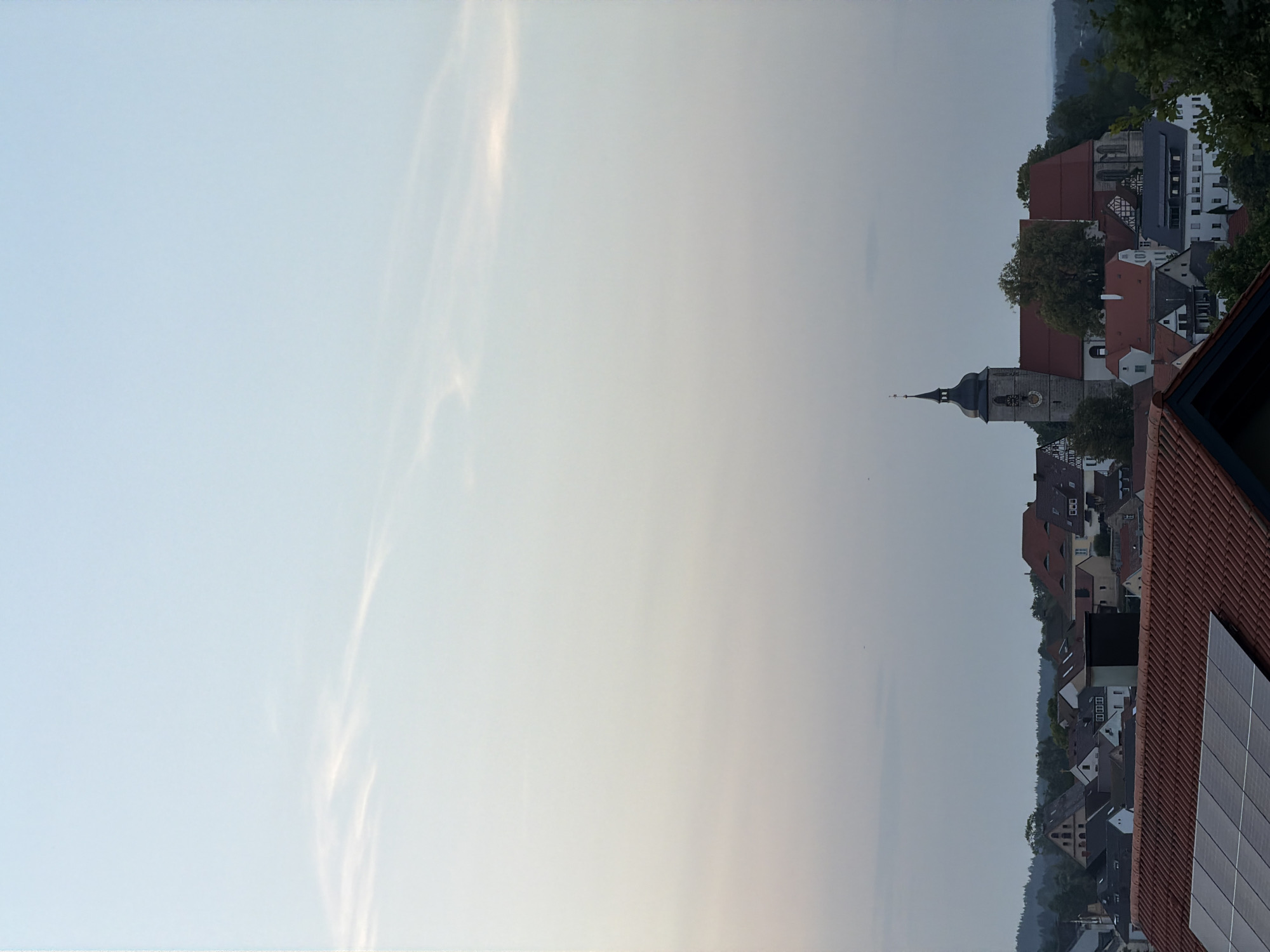 The image shows a panoramic view of the town of Creußen, Germany, at dusk. The sky is a soft gradient of blue and pale grey, with wispy clouds. In the foreground, a red-tiled roof with solar panels is visible. The middle ground features a cluster of buildings, including a prominent church with a tall, dark spire. The church is made of stone and has a clock face. Other buildings in the town have red-tiled roofs and a mix of architectural styles. Trees and greenery are interspersed among the buildings, and a line of dark trees forms a backdrop in the distance. The overall impression is of a peaceful, historic European town as day transitions to night.