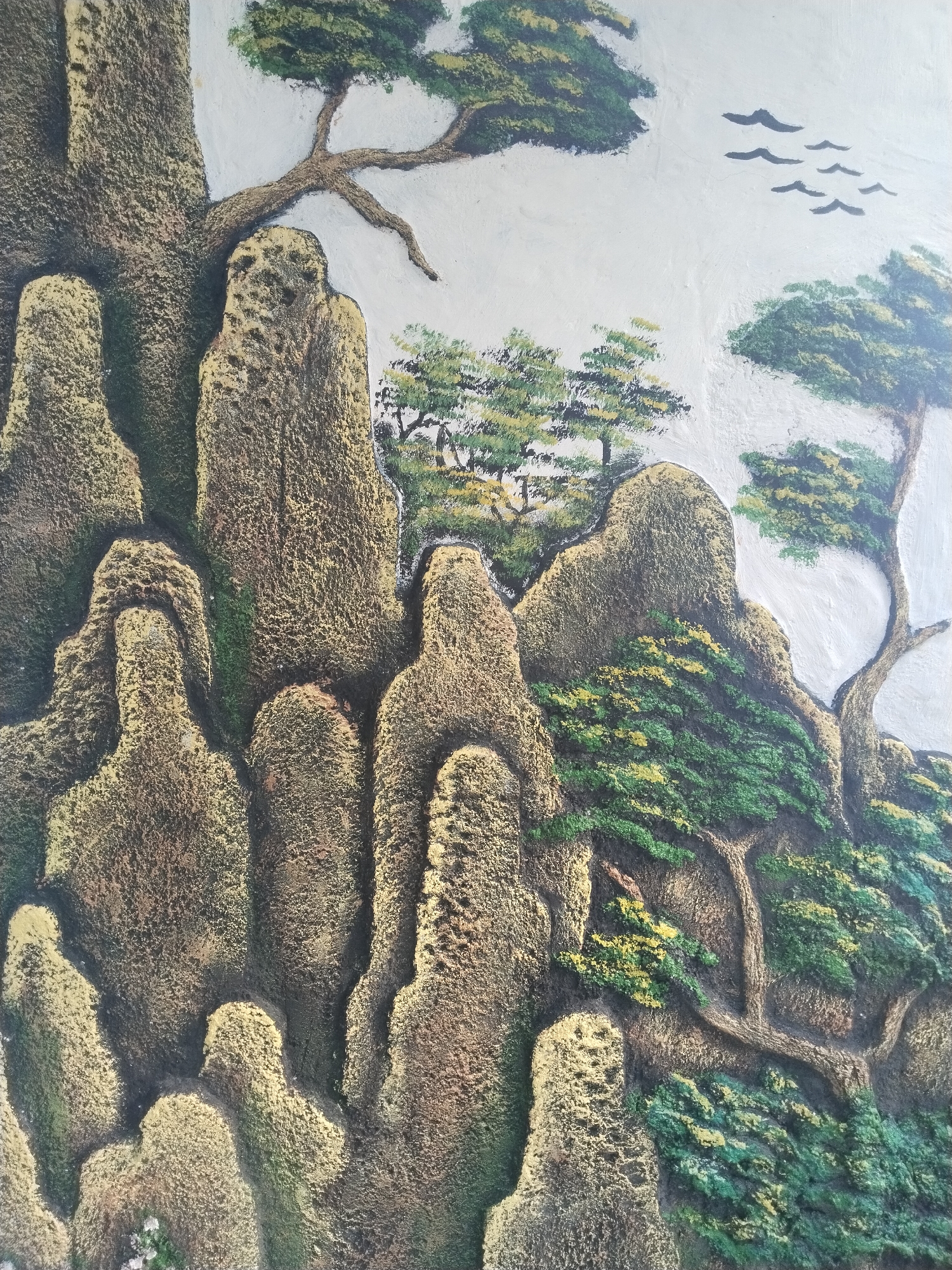 This media file showcases a vibrant, textured relief sculpture or mural depicting a stylized mountainous landscape. The art features prominent, rugged rock formations that dominate the scene, rendered in earthy tones of brown, gold, and dark green, giving them a mossy, aged appearance. The textured surface of the relief impressively mimics the rough, craggy nature of rock and the bark of trees.
Interspersed among the tall, vertical rock peaks are numerous trees. These trees, some resembling pines with their distinct branch structures, are painted in various shades of green, from dark forest green to lighter, almost yellowish-green for the foliage, suggesting highlights or different species. The branches themselves are sculpted in relief, adding to the three-dimensional quality of the artwork.
The upper section of the artwork depicts a clear, bright sky, painted in a light, almost white hue. A small flock of dark, silhouetted birds is visible flying across this expansive sky in the upper right portion of the image, adding a sense of scale and movement to the otherwise static natural scene.
The overall artistic style strongly evokes traditional East Asian landscape paintings, particularly 'shan shui' (mountain-water) art, characterized by its emphasis on mountains, trees, and ethereal compositions. This detailed and tactile artwork is located in Bangsri, Indonesia. There is no visible text in the image. The time of day appears to be bright daytime, under clear skies.