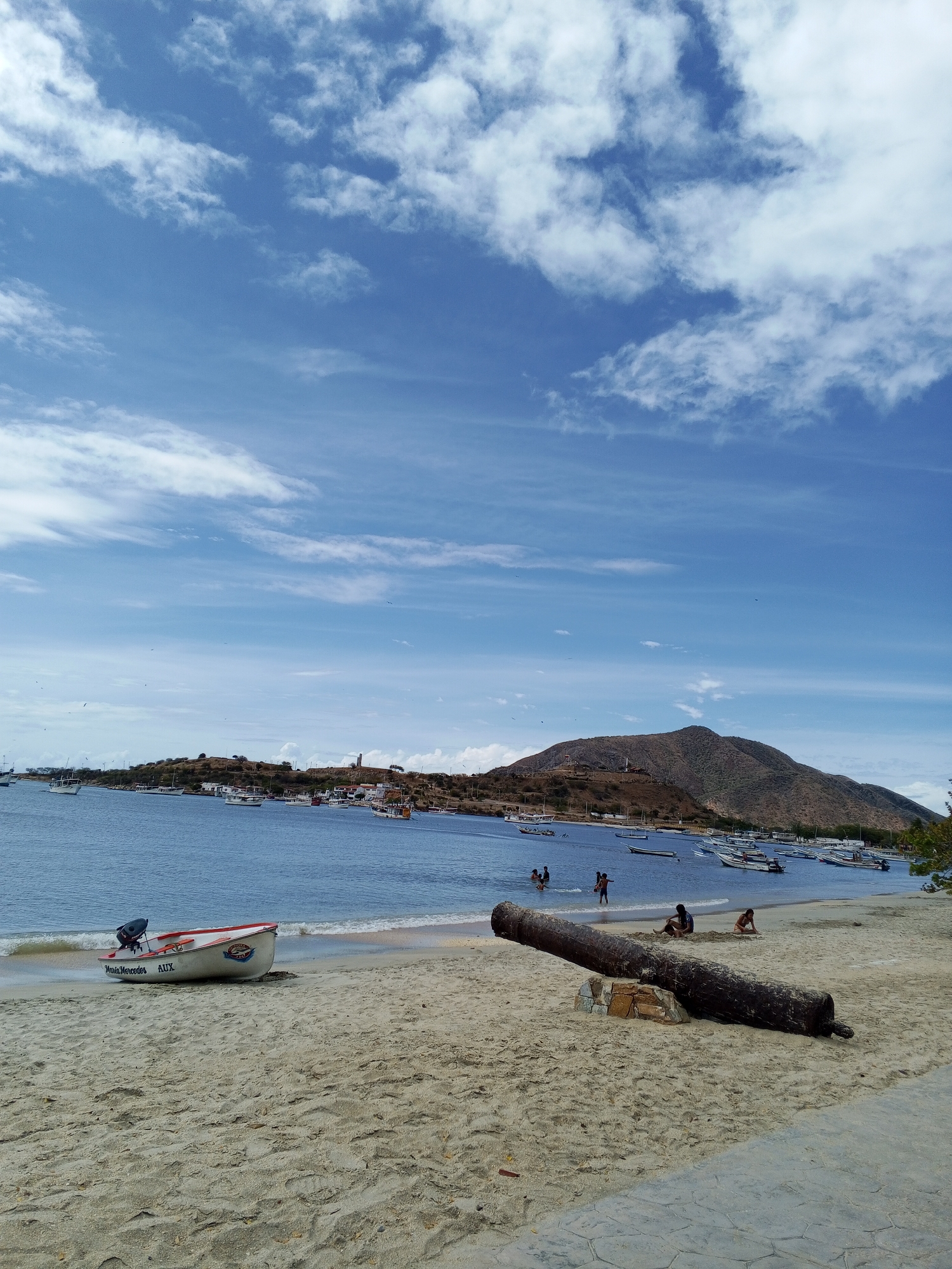 The image shows a beach scene in the city of Unknown, Unknown. In the foreground, there's a sandy beach with a small white boat on the left and an old, large, rusty cannon on the right, mounted on stones. Several people are scattered along the shoreline; some are in the water and others are sitting on the sand. The ocean fills the midground, with numerous boats anchored in the bay. A landmass with a mountain and some buildings is visible in the background, under a sky dotted with clouds. The day appears sunny.