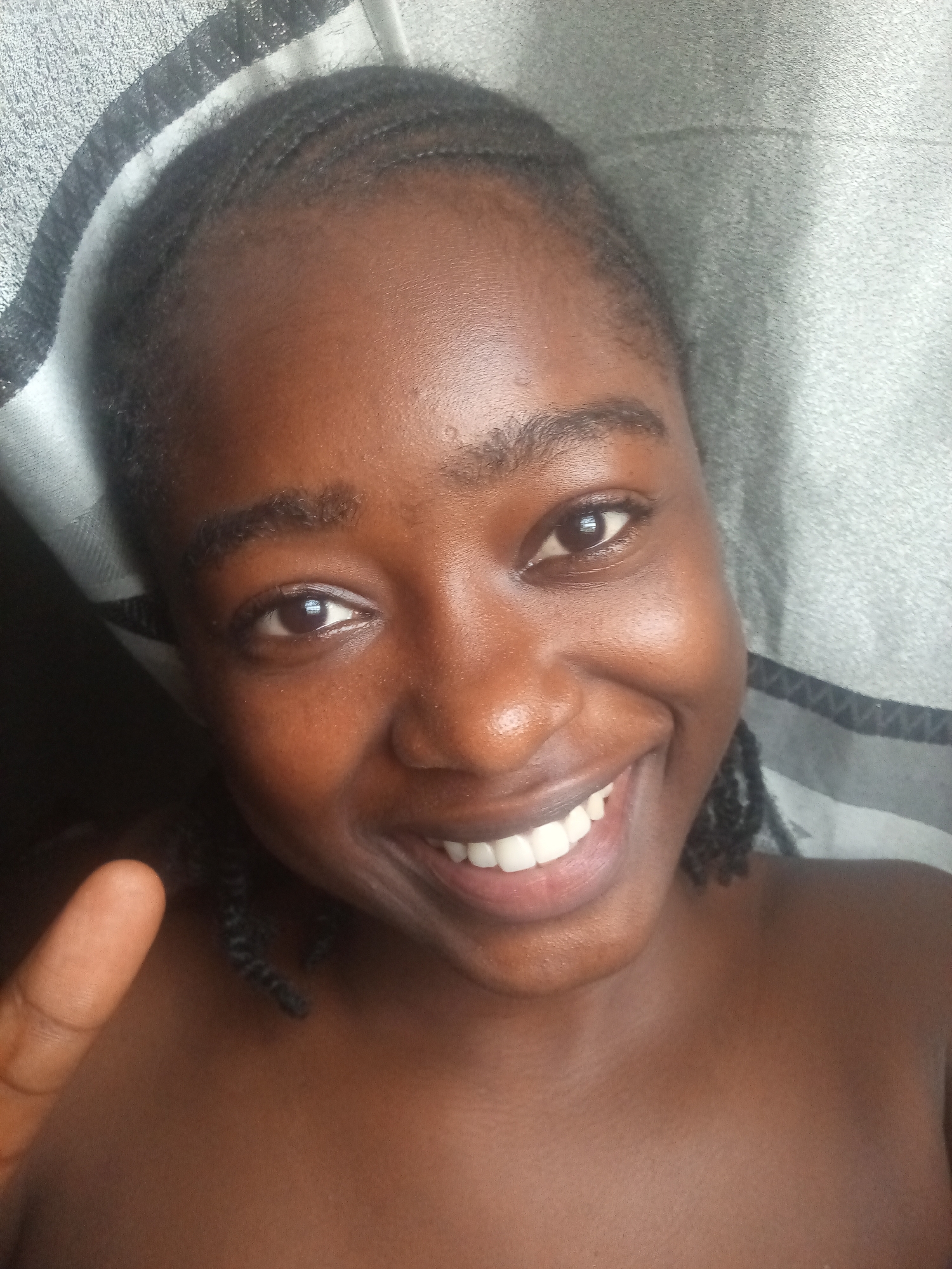 A young Black female, who appears to be in her late teens or early twenties, is shown in a close-up, head-and-shoulders selfie. She has dark skin and her hair is styled in small braids that are pulled back from her face. She is smiling broadly, revealing white teeth. Her eyes are dark brown. A portion of her left thumb is visible in the lower left corner of the image, held up in a peace sign gesture. She is wearing a dark top, which exposes her shoulders and upper chest. The background is a textured gray surface, possibly a blanket or towel. The lighting is soft and diffused. The image quality is average, with some graininess. There are no discernible landmarks or contextual clues to indicate a specific location in Rafin Par, Nigeria.