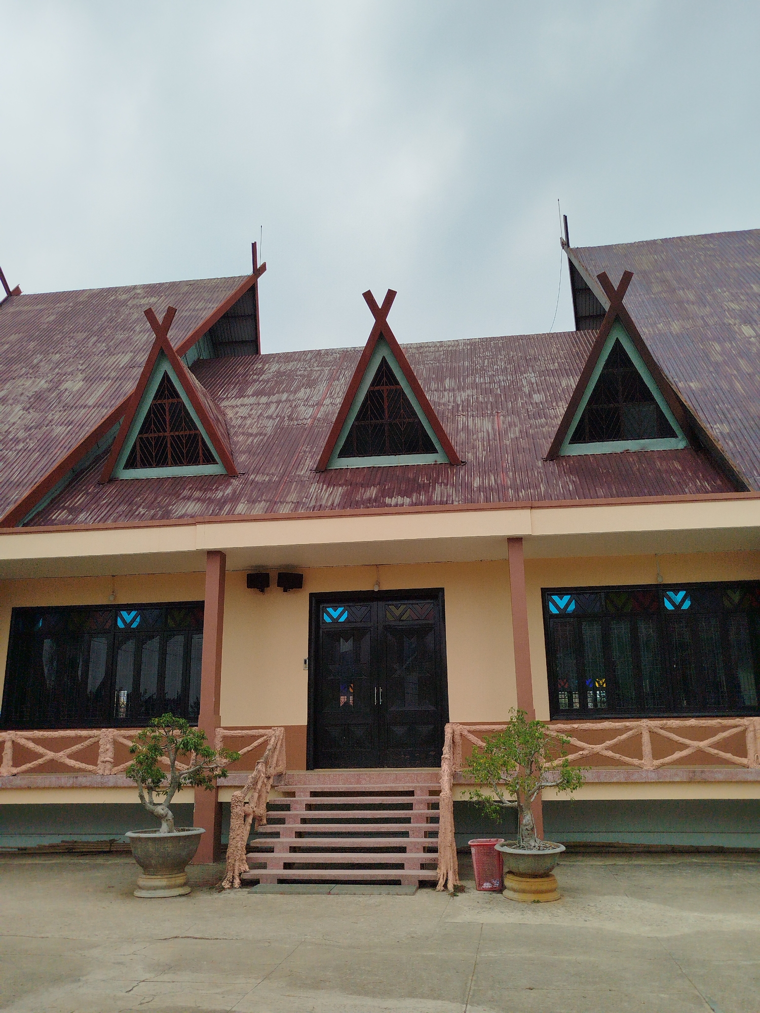 A daytime shot of a two-story building in K'Long, Vietnam. The building has a rusty red corrugated iron roof with three triangular dormers framed in green. The main structure is light beige with dark brown pillars and window frames. Two black double doors serve as the entrance, approached by a series of pink-ish beige steps with similarly colored railings mimicking natural tree branches. Two bonsai trees in large stone pots flank the steps. The sky is overcast, and the ground is a gray concrete slab.