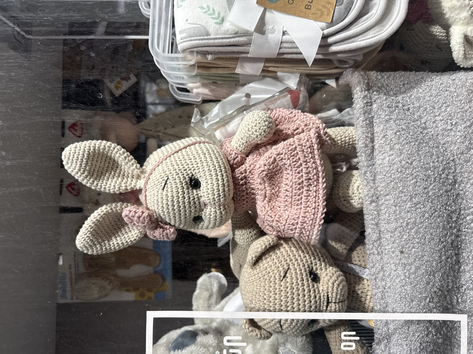 The image shows a display of handmade crochet toys, including a bunny and a bear, inside a shop window. The bunny is wearing a pink dress and a pink bow on its head. The bear is a light brown color. Behind the toys, there are other baby items, such as blankets and bibs, hanging on hangers. The shop appears to be located in Yerevan, Armenia, as indicated by the Cyrillic text visible on the price tags and packaging. The window is dirty, with streaks and smudges obscuring some of the view. The lighting suggests it is daytime.
